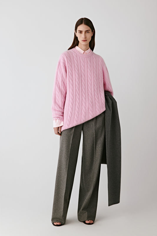 Front view of the pink cable-knit cashmere, alpaca and silk pullover, styled with wide grey trousers.