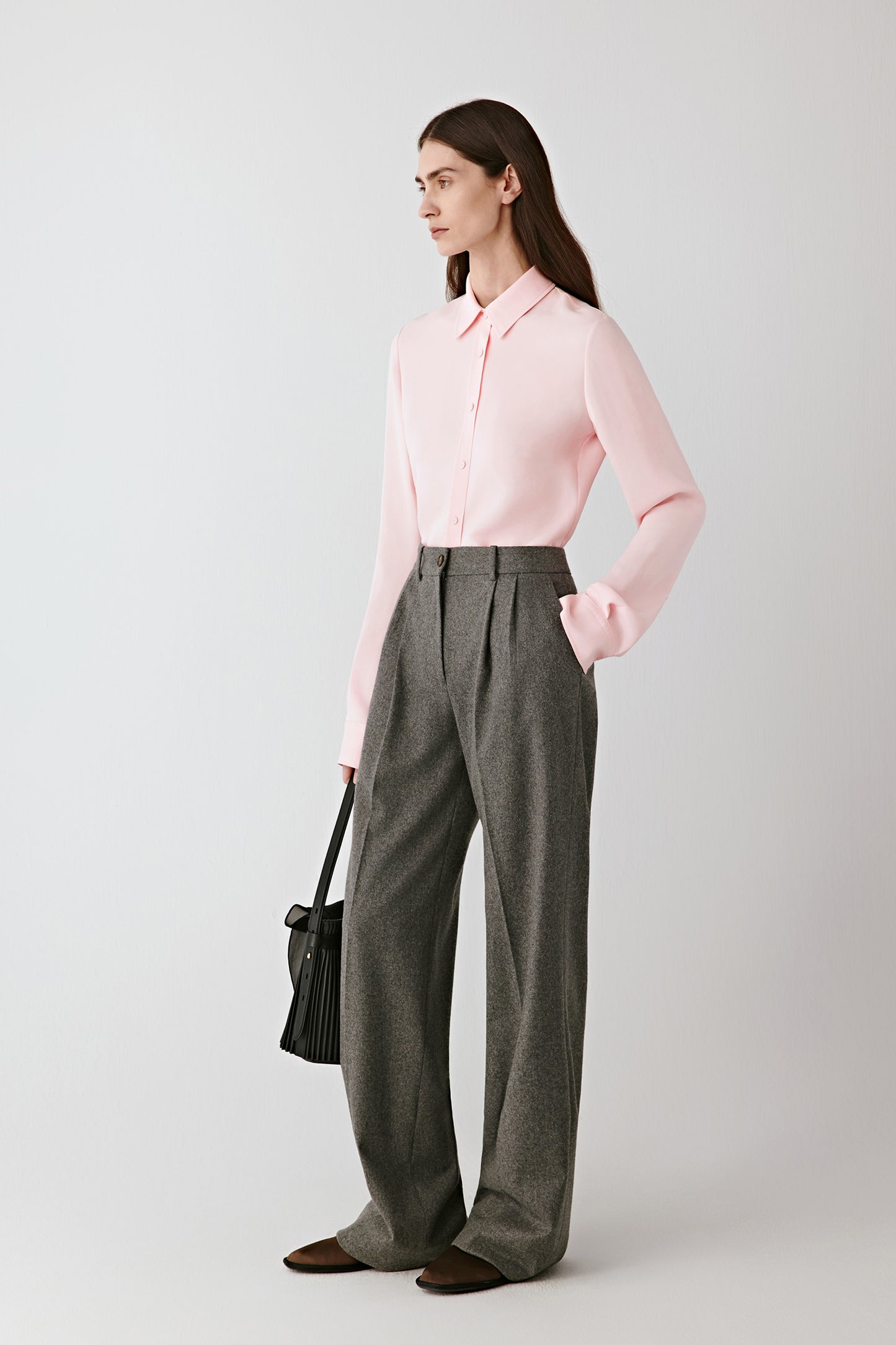 Full-length front view of the model wearing the pale pink silk crêpe shirt tucked into wide grey trousers, showing the shirt’s fluid drape and subtle sheen under soft light.