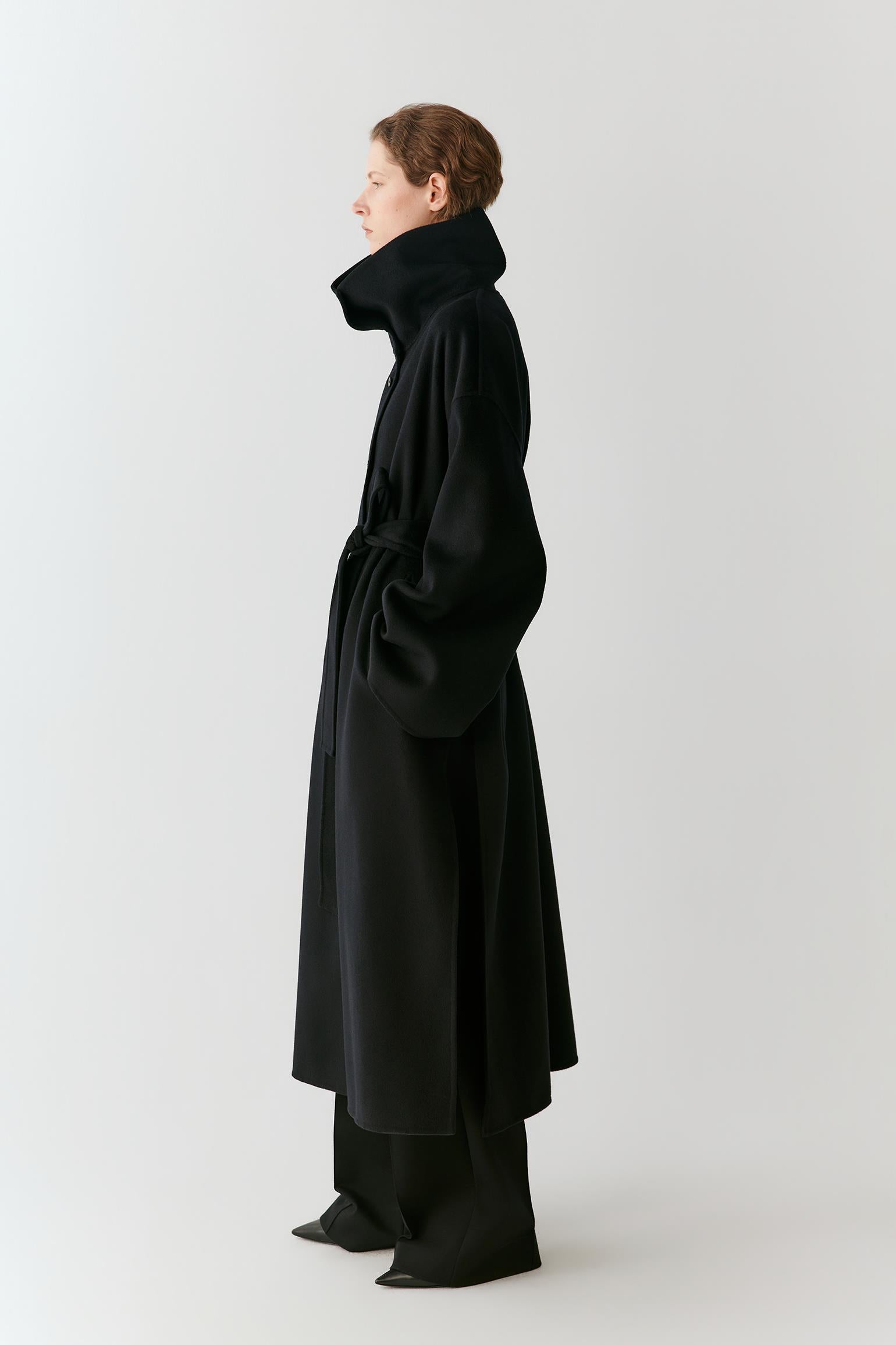 A model in profile wearing a long black coat with a high structured collar, matching wide-leg trousers, and pointed shoes, photographed against a plain light gray studio background.