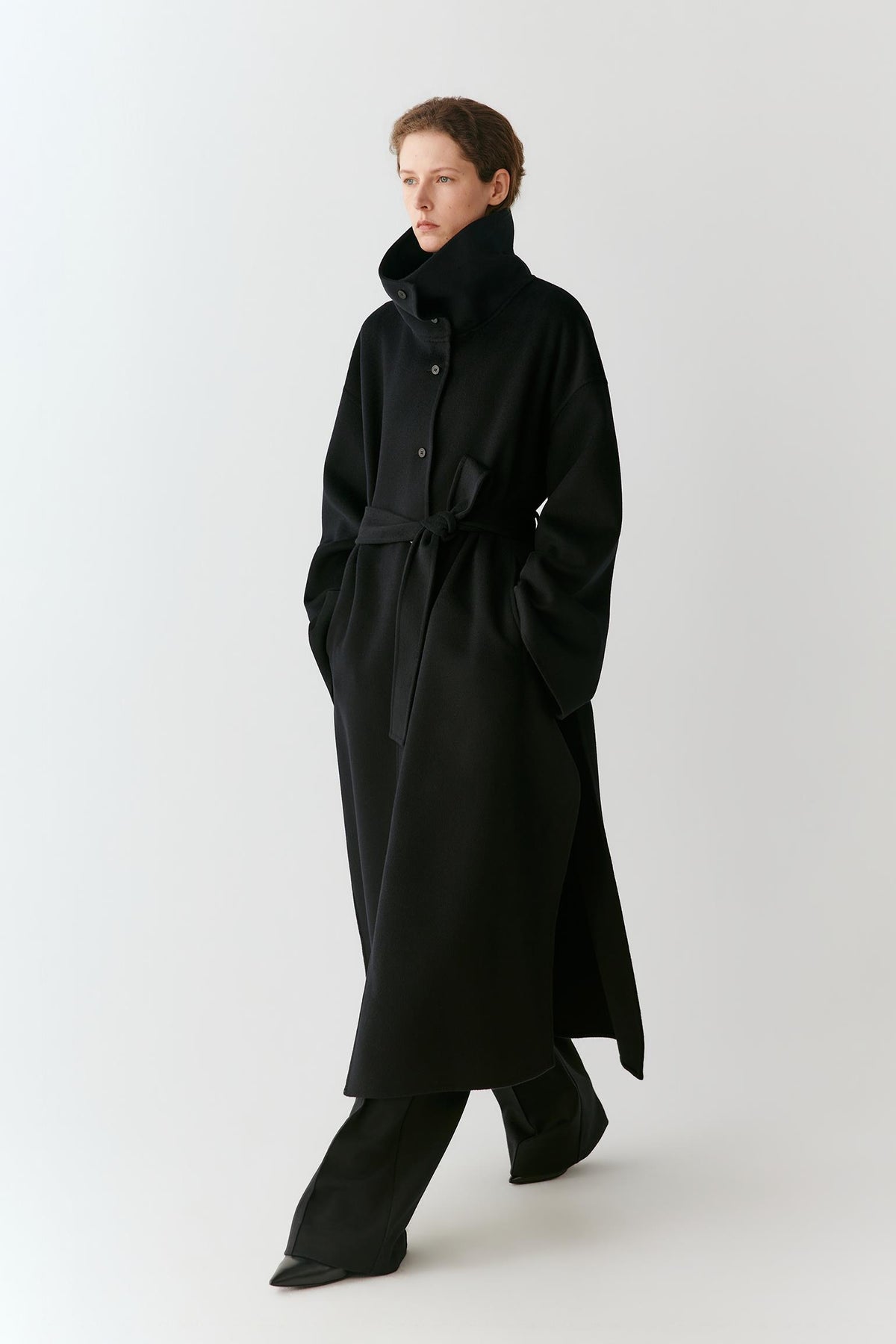 A model wearing a long black coat with a high collar and waist tie, paired with black trousers, photographed against a plain light gray studio background.