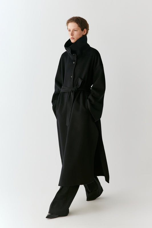 A model wearing a long black coat with a high collar and waist tie, paired with black trousers, photographed against a plain light gray studio background.