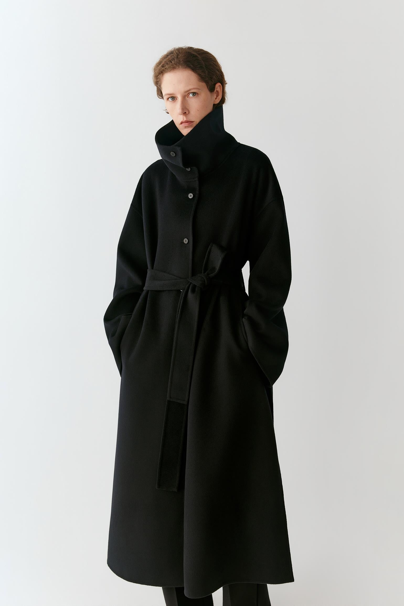 A model wearing a long black wool coat with a high collar, button front, and matching waist tie, photographed against a plain light gray studio background.