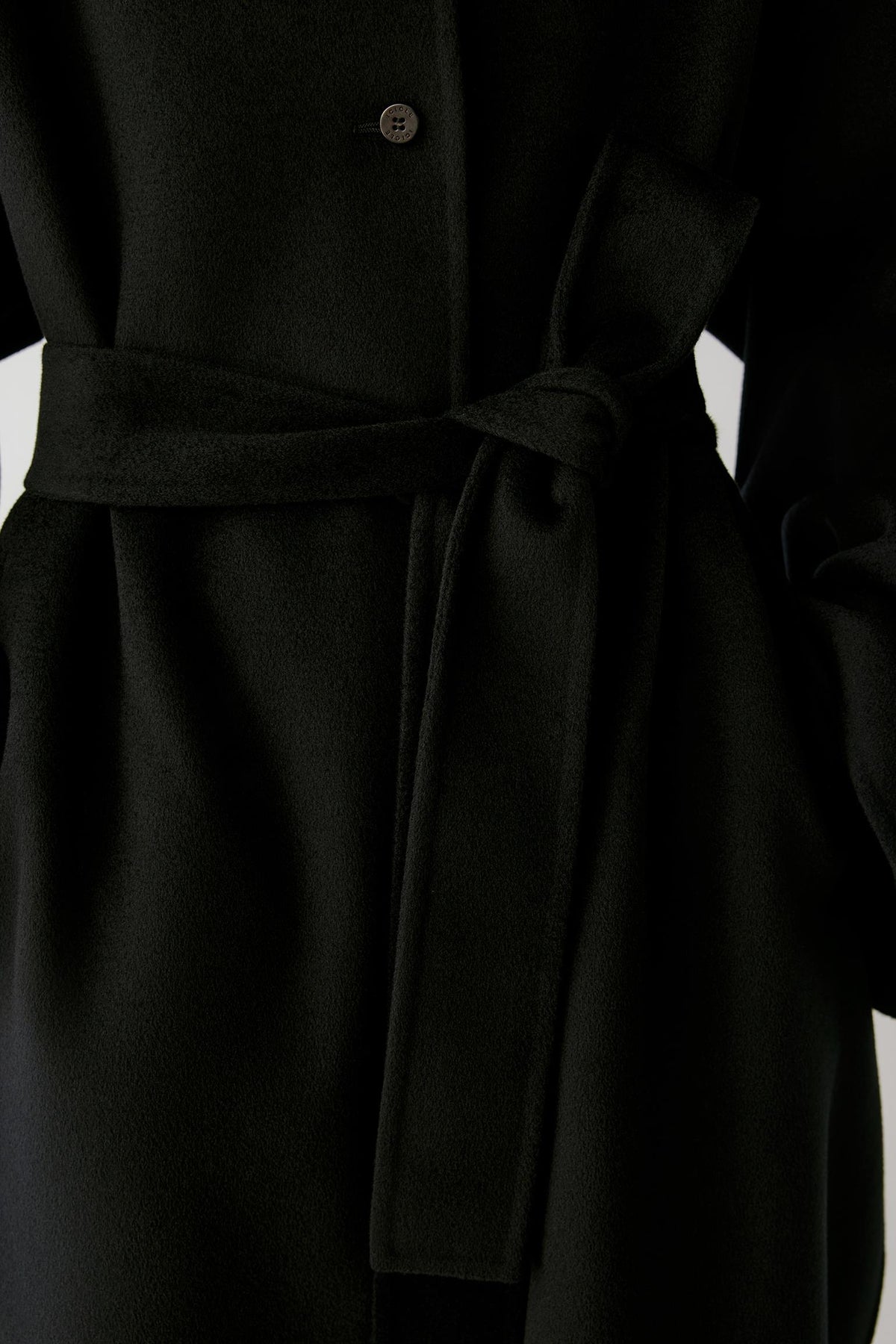 Close-up of a black belted coat with a single visible button and soft fabric folds, photographed against a dark background.