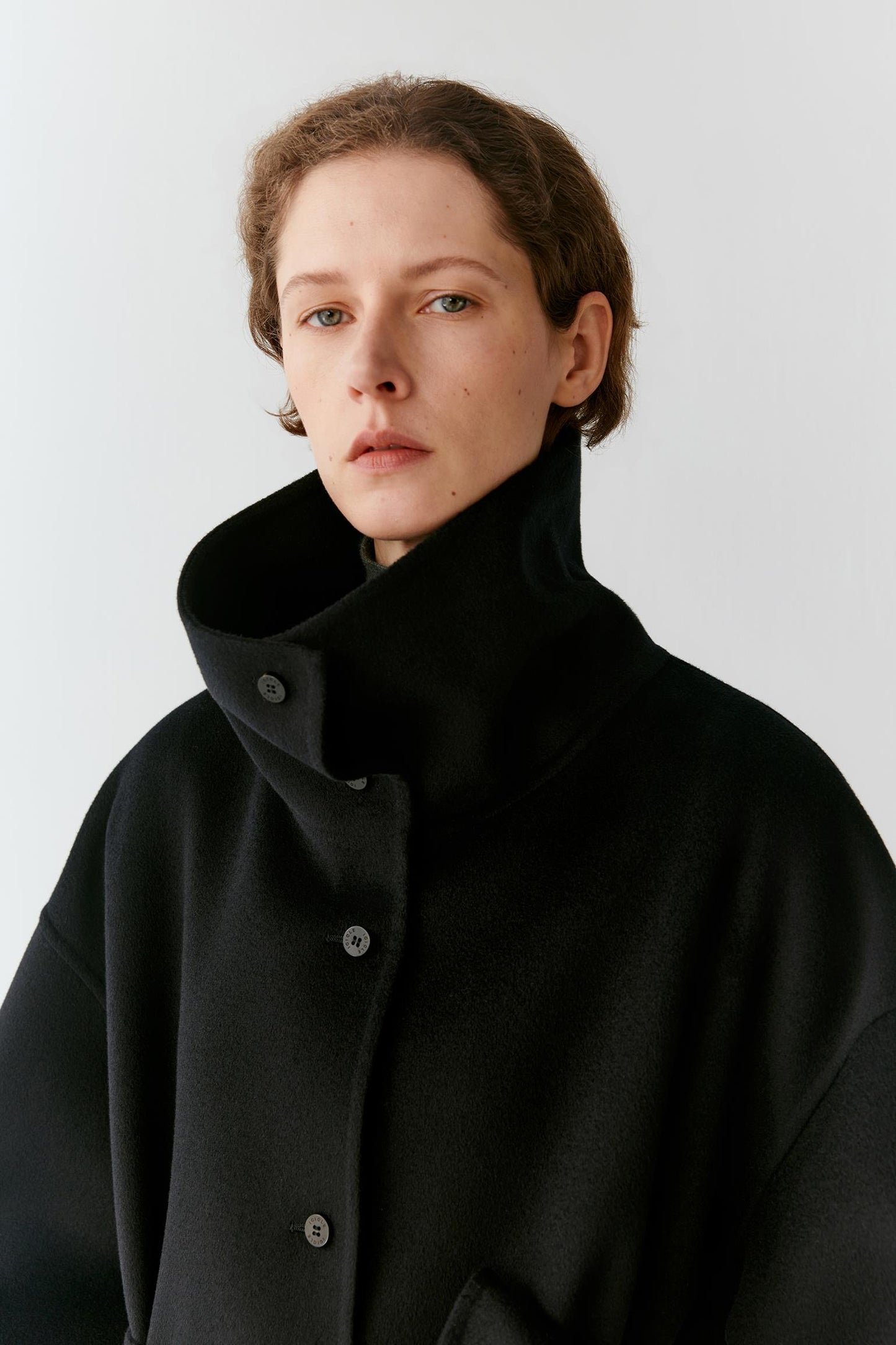 A model wearing a black oversized coat with a very high structured collar and dark buttons, photographed against a plain light gray studio background.