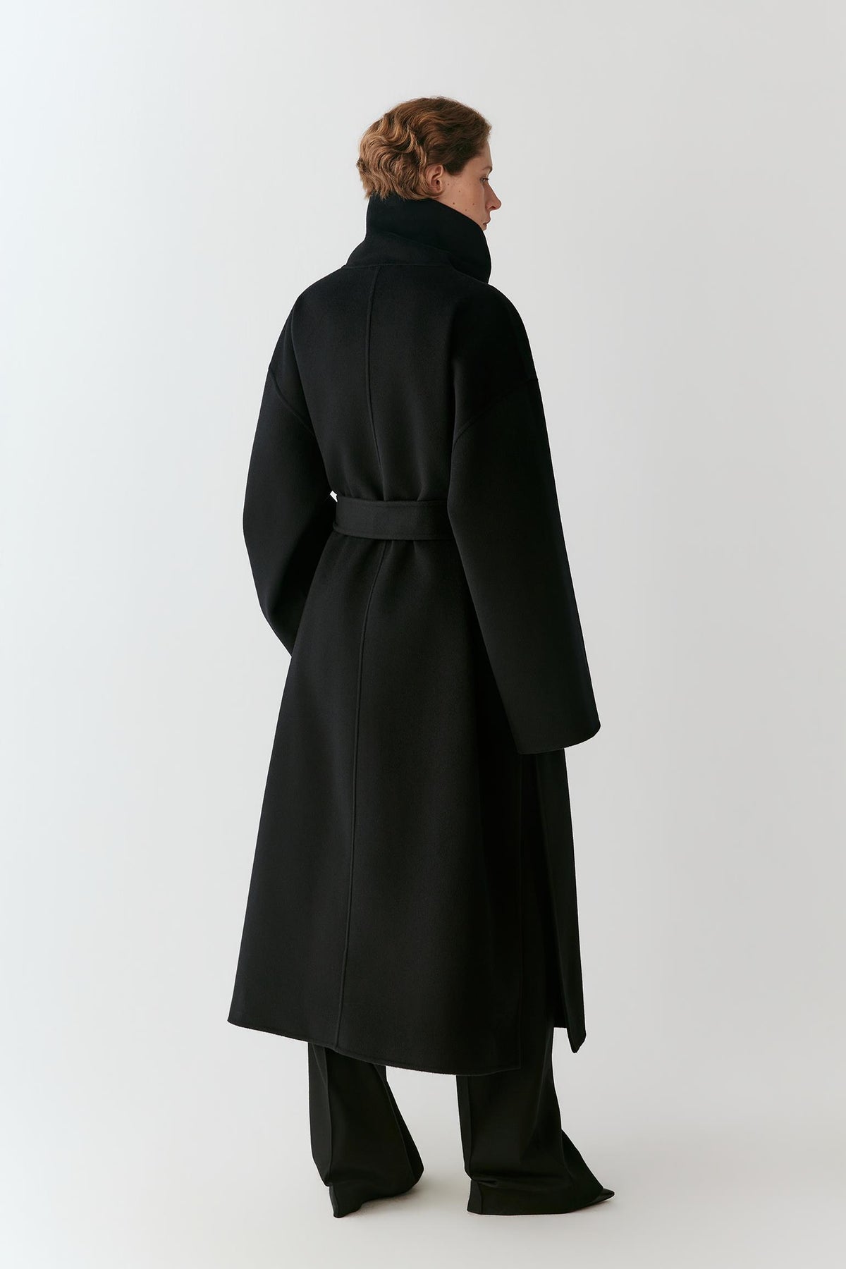 A model shown from the back wearing a long black wool coat with a high collar and waist belt, paired with wide-leg trousers, against a light gray studio background.