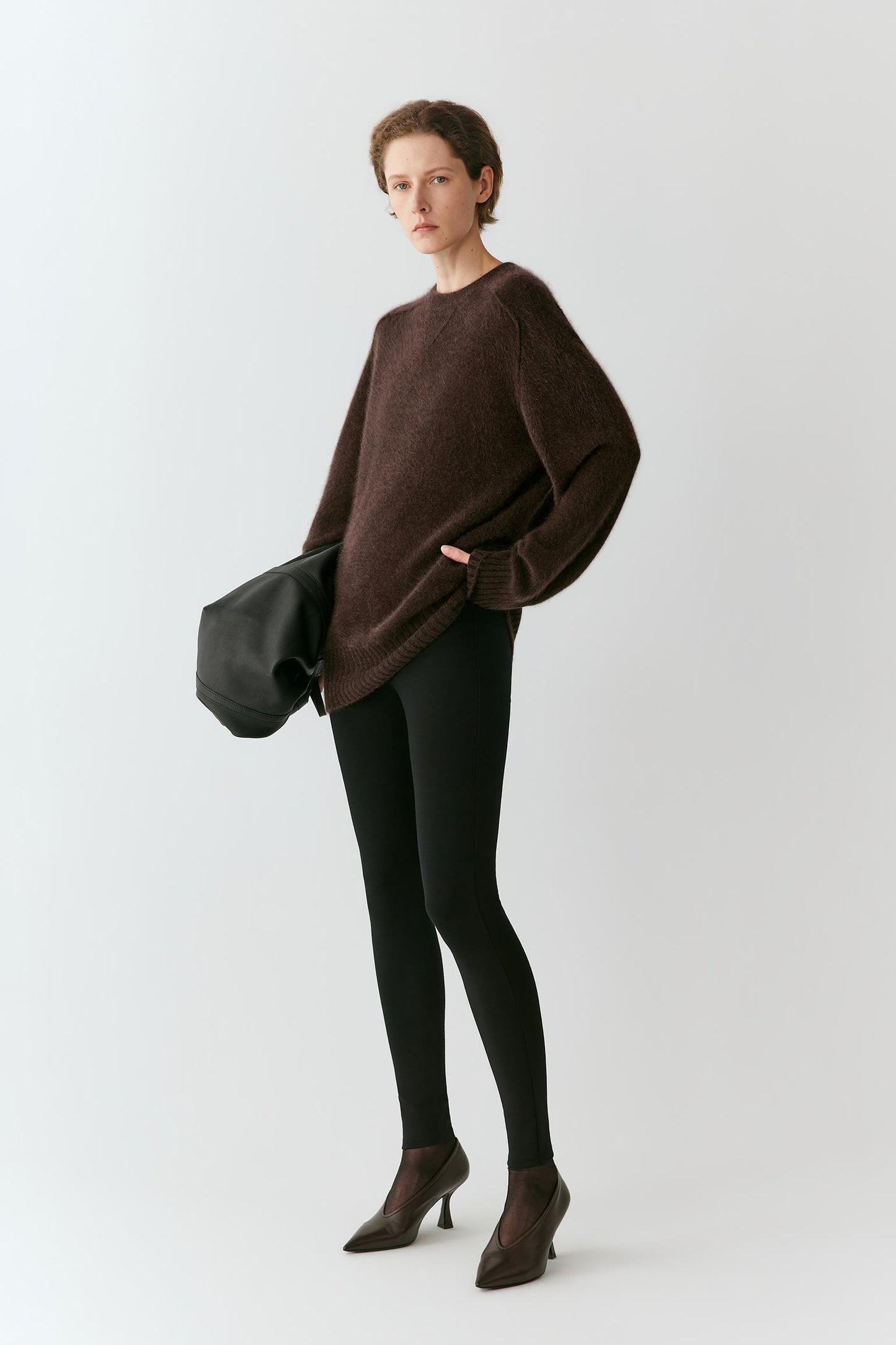 A model stands against a light gray studio background wearing an oversized dark brown knit sweater, black leggings, dark brown pointed-toe heels, and holding a black clutch bag.