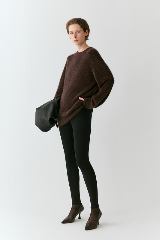 A model stands against a light gray studio background wearing an oversized dark brown knit sweater, black leggings, dark brown pointed-toe heels, and holding a black clutch bag.