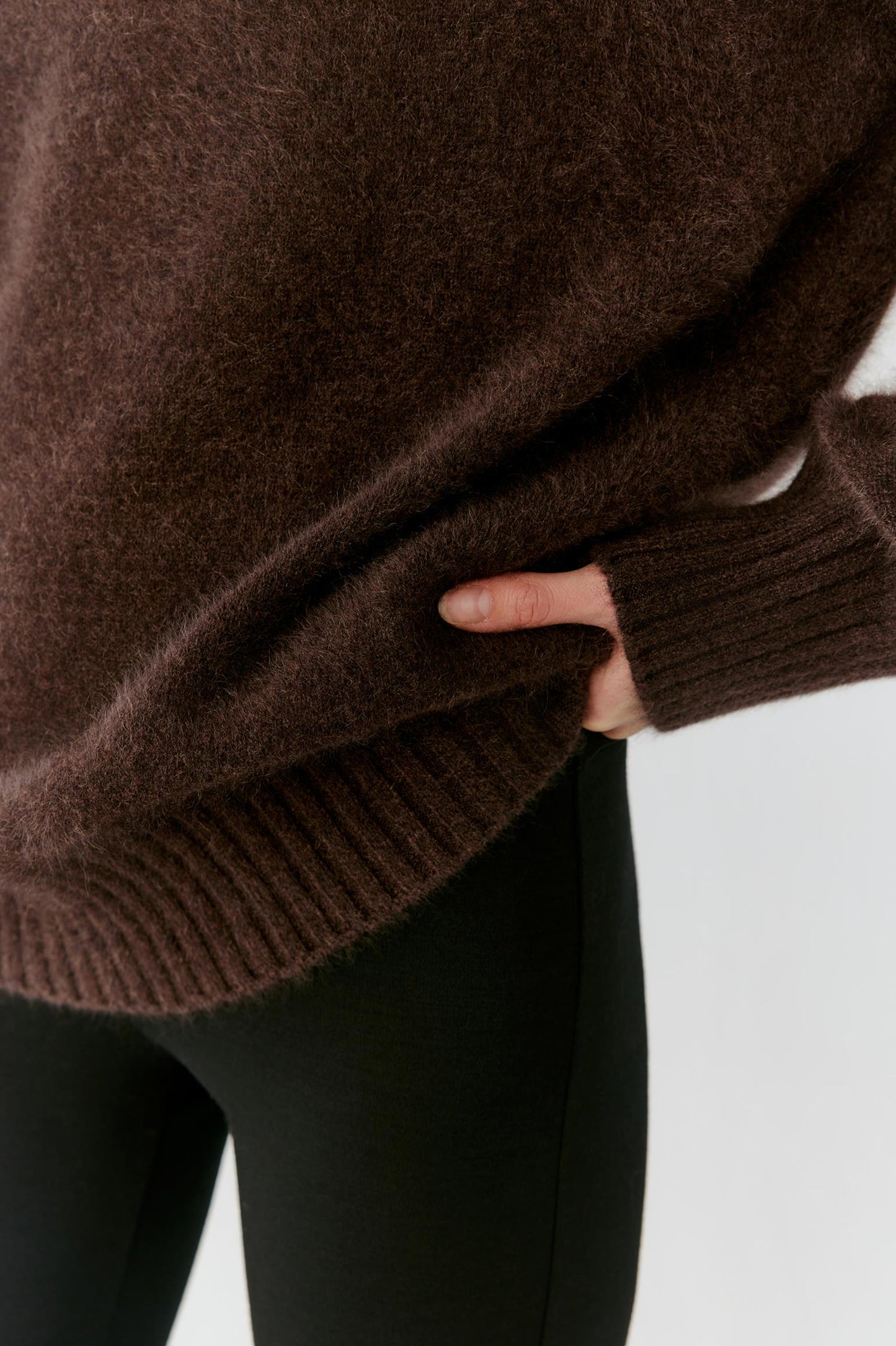 Close-up of a person wearing an oversized dark brown fuzzy knit sweater with ribbed cuffs and hem, paired with black leggings, against a light gray background.