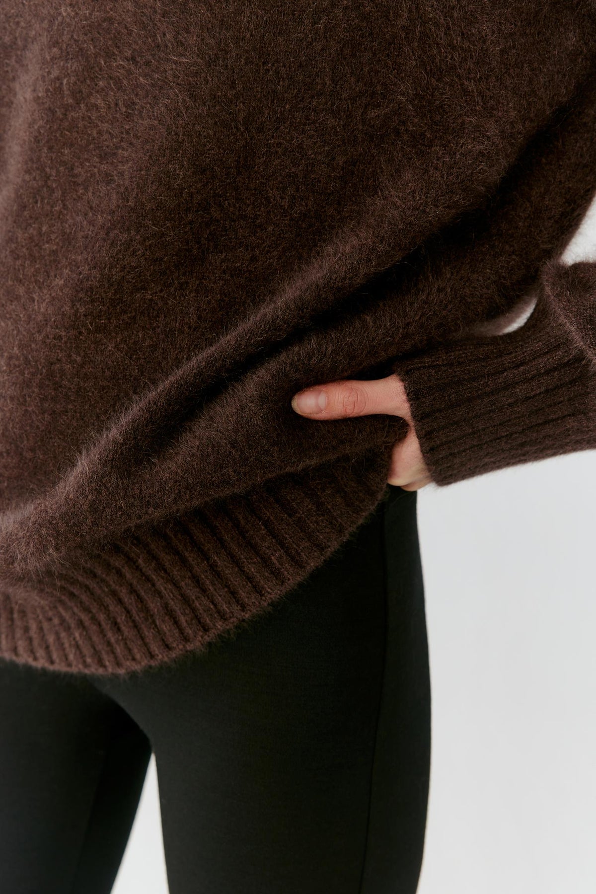 Close-up of a person wearing an oversized dark brown fuzzy knit sweater with ribbed cuffs and hem, paired with black leggings, against a light gray background.