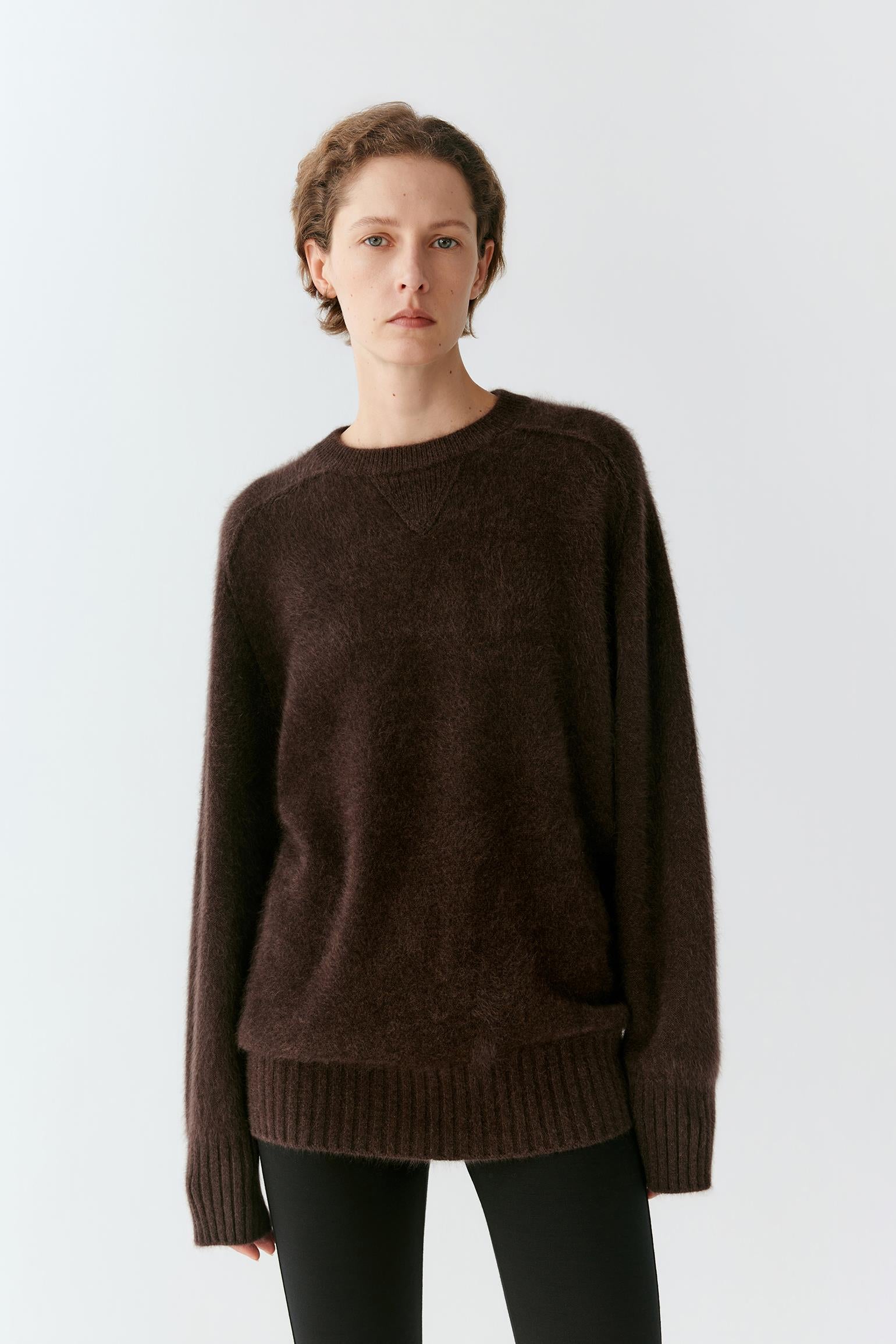 A woman wearing a dark brown fuzzy knit crewneck sweater and black pants, standing against a light gray studio background.