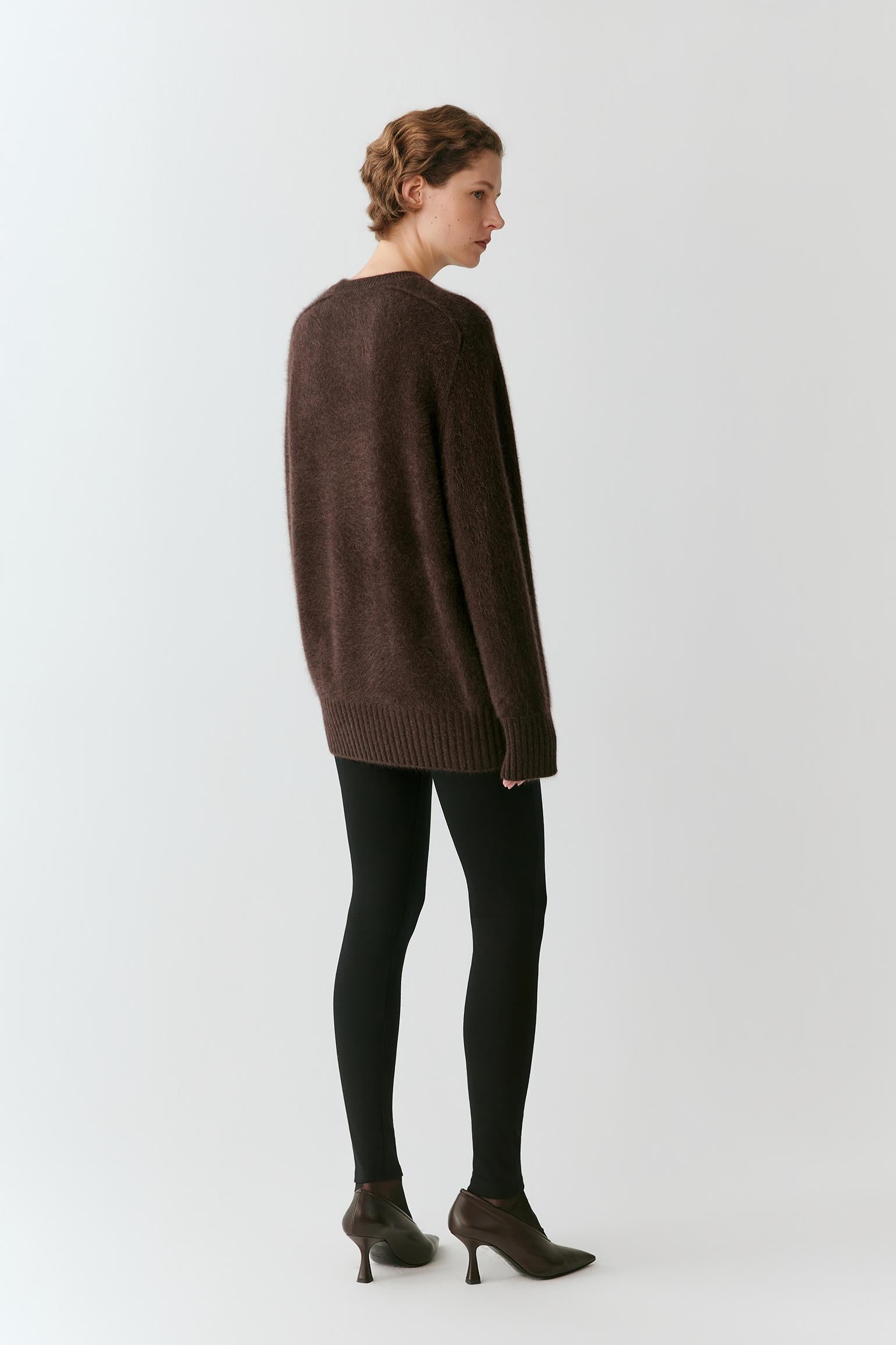 A woman stands in profile wearing an oversized dark brown knit sweater, black tights, and dark brown heeled ankle boots against a plain light gray studio background.