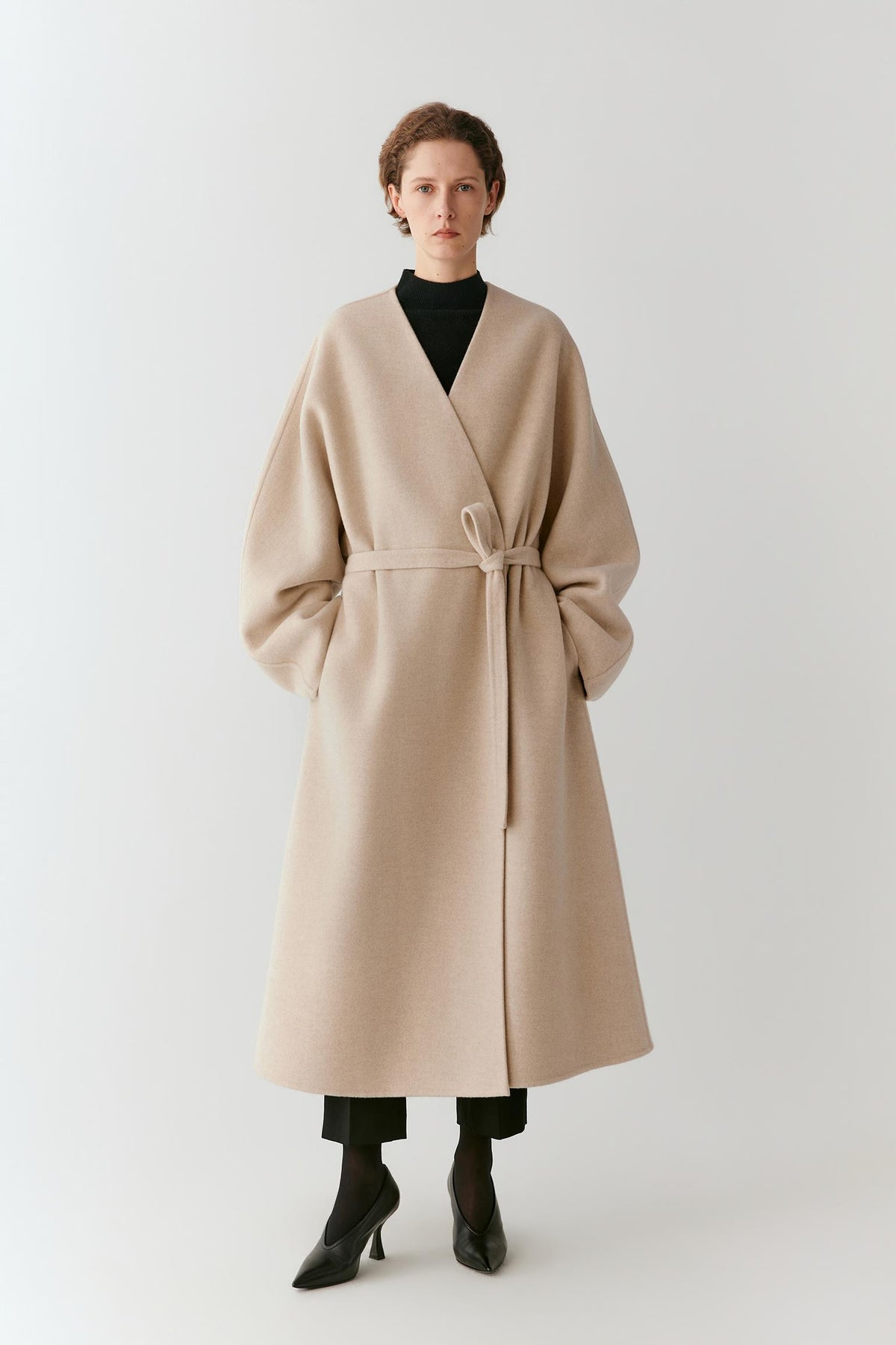Front view of a woman wearing a beige melange wool and cashmere coat, showing the long wrap silhouette, V-neckline and self-tie belt.