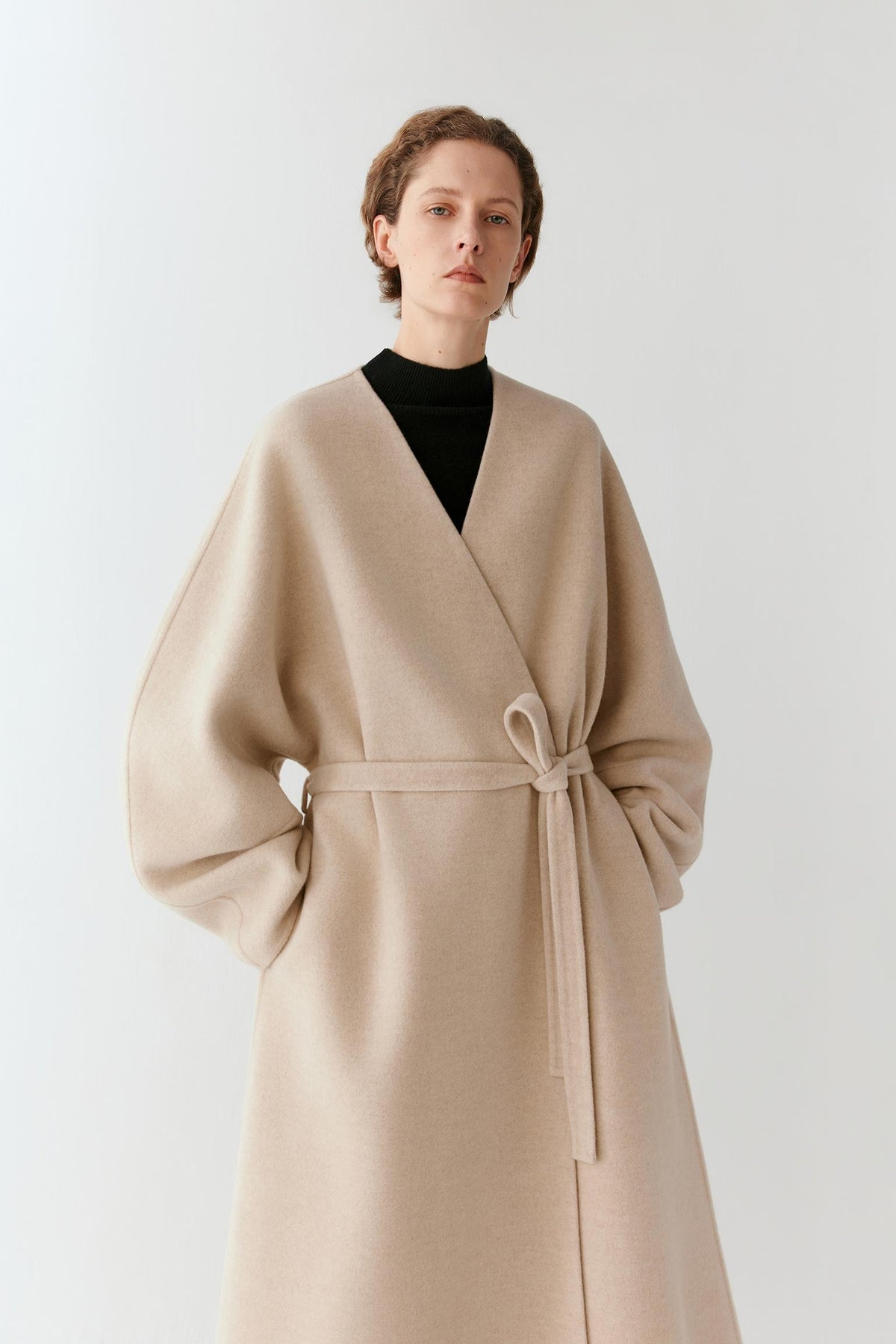 Three-quarter view highlighting the wrap-front construction, self-tie waist and fluid drape of the beige melange coat.