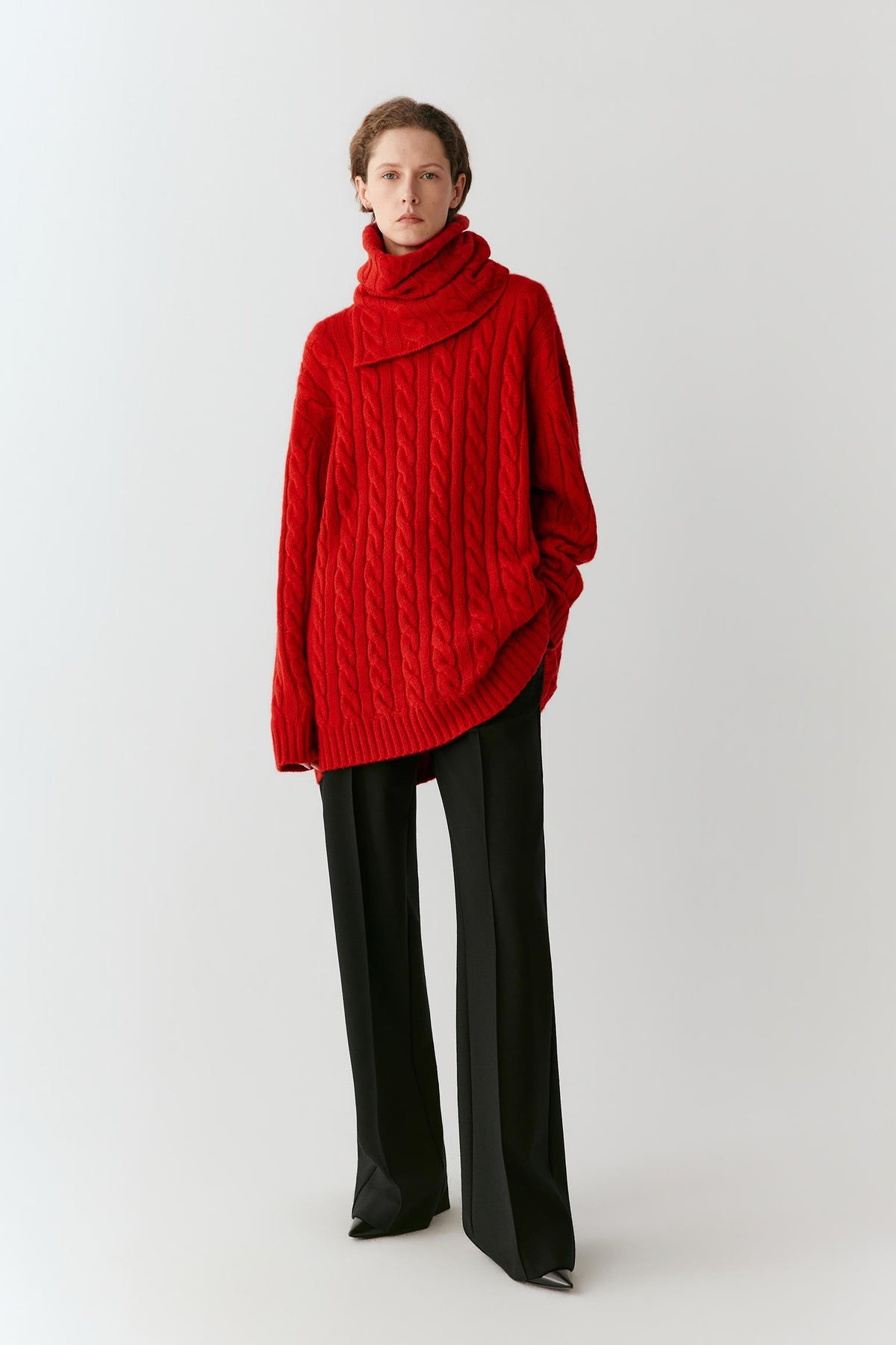 A model wearing a vibrant red cable-knit cowl-neck sweater and black wide-leg trousers, standing against a plain light gray studio background.