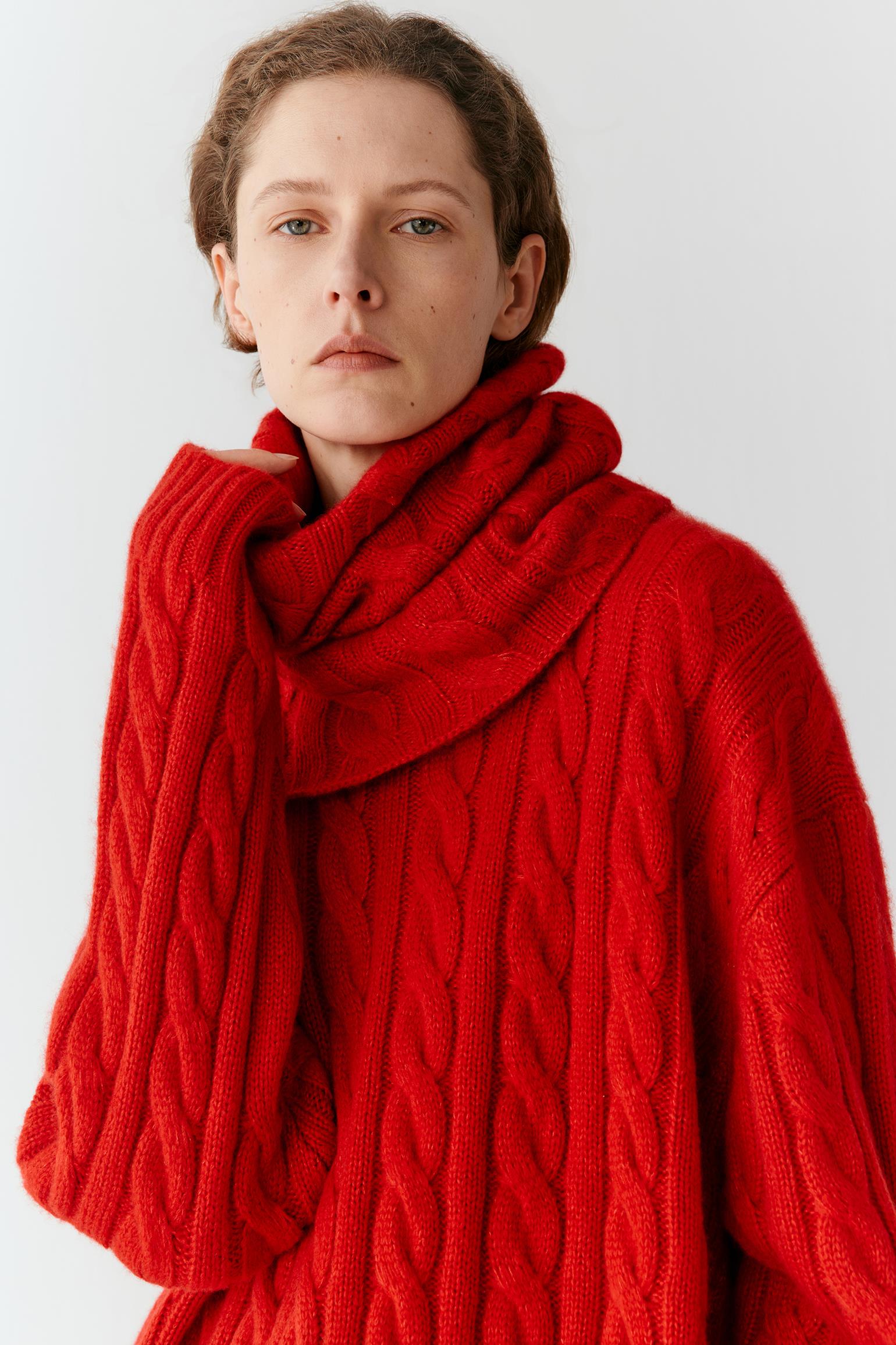 A close-up of a model wearing a vibrant red cable-knit sweater and matching thick red cowl scarf against a plain light gray background.