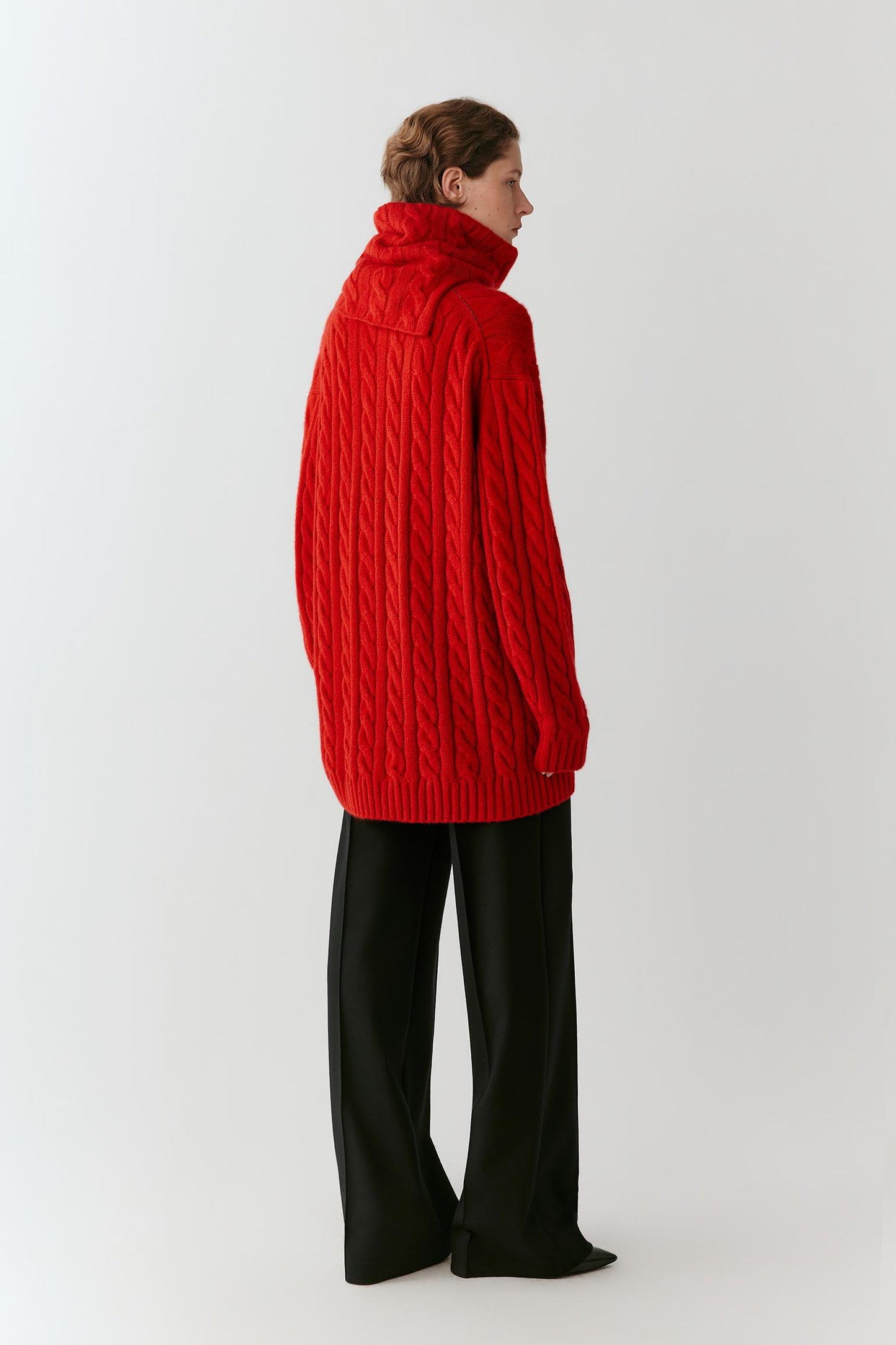 A model shown from the back wearing a bright red cable-knit mock-neck sweater and black wide-leg trousers, standing against a plain light gray studio background.