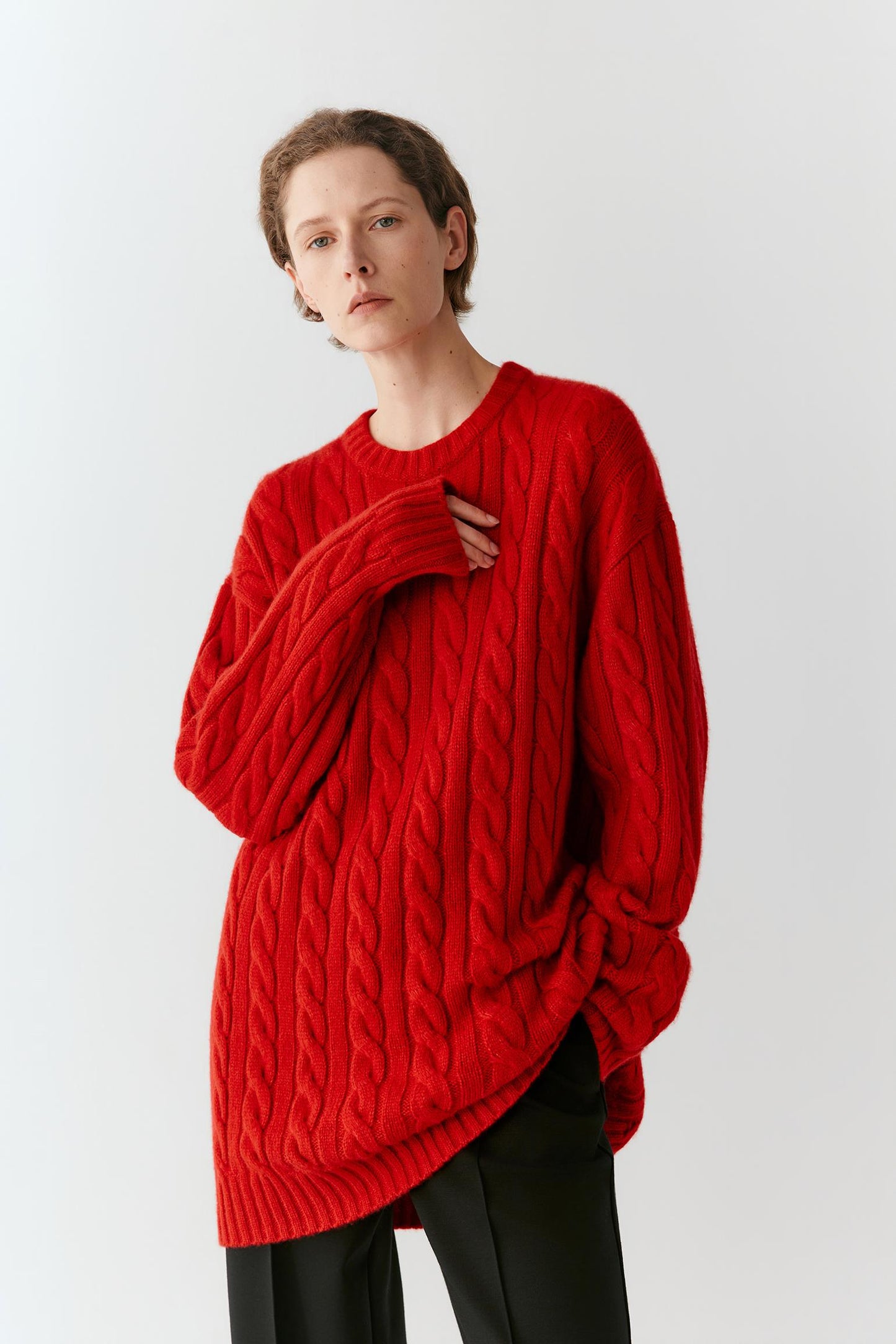 A model wearing a vibrant red cable-knit sweater and black trousers, standing against a plain light gray studio background.