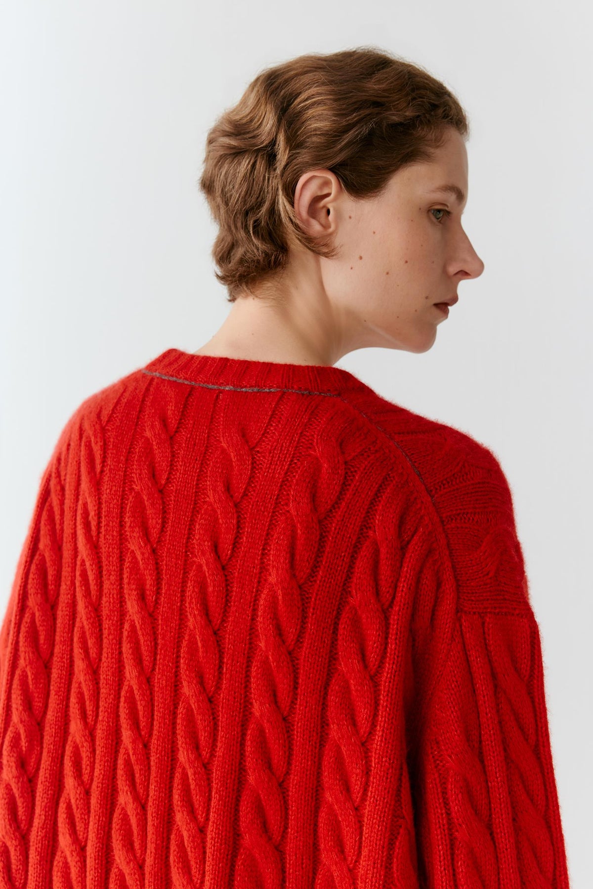 A woman shown from the back and side wearing a bright red cable-knit sweater against a plain light gray studio background.