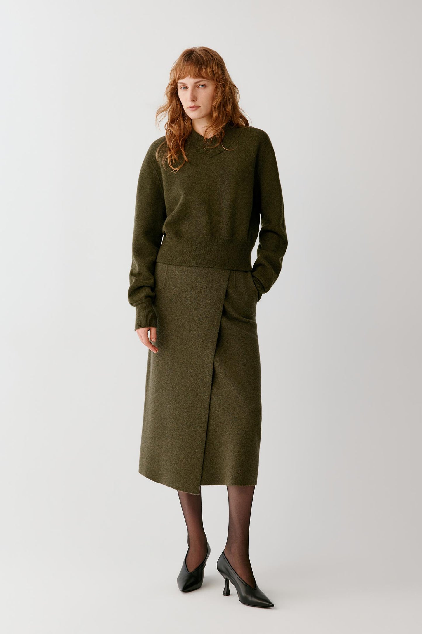 A model with red hair stands against a light gray studio background wearing an olive green knit sweater and matching olive green wrap skirt, paired with black tights and black pointed-toe heels.