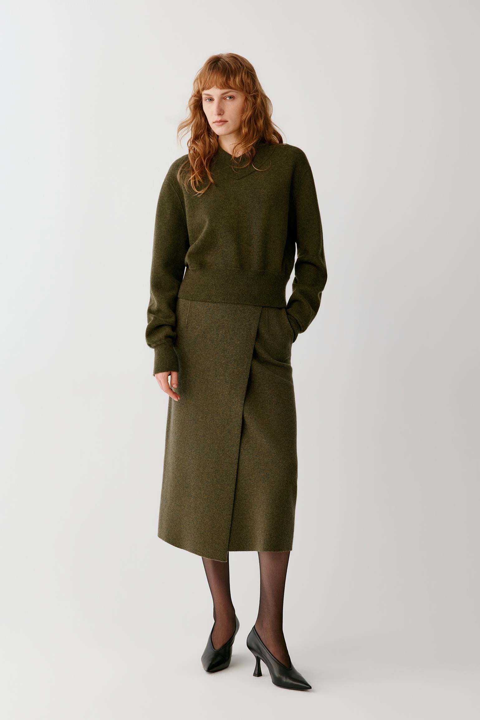 A model with red hair stands against a light gray studio background wearing an olive green knit sweater and matching olive green wrap skirt, paired with black tights and black pointed-toe heels.