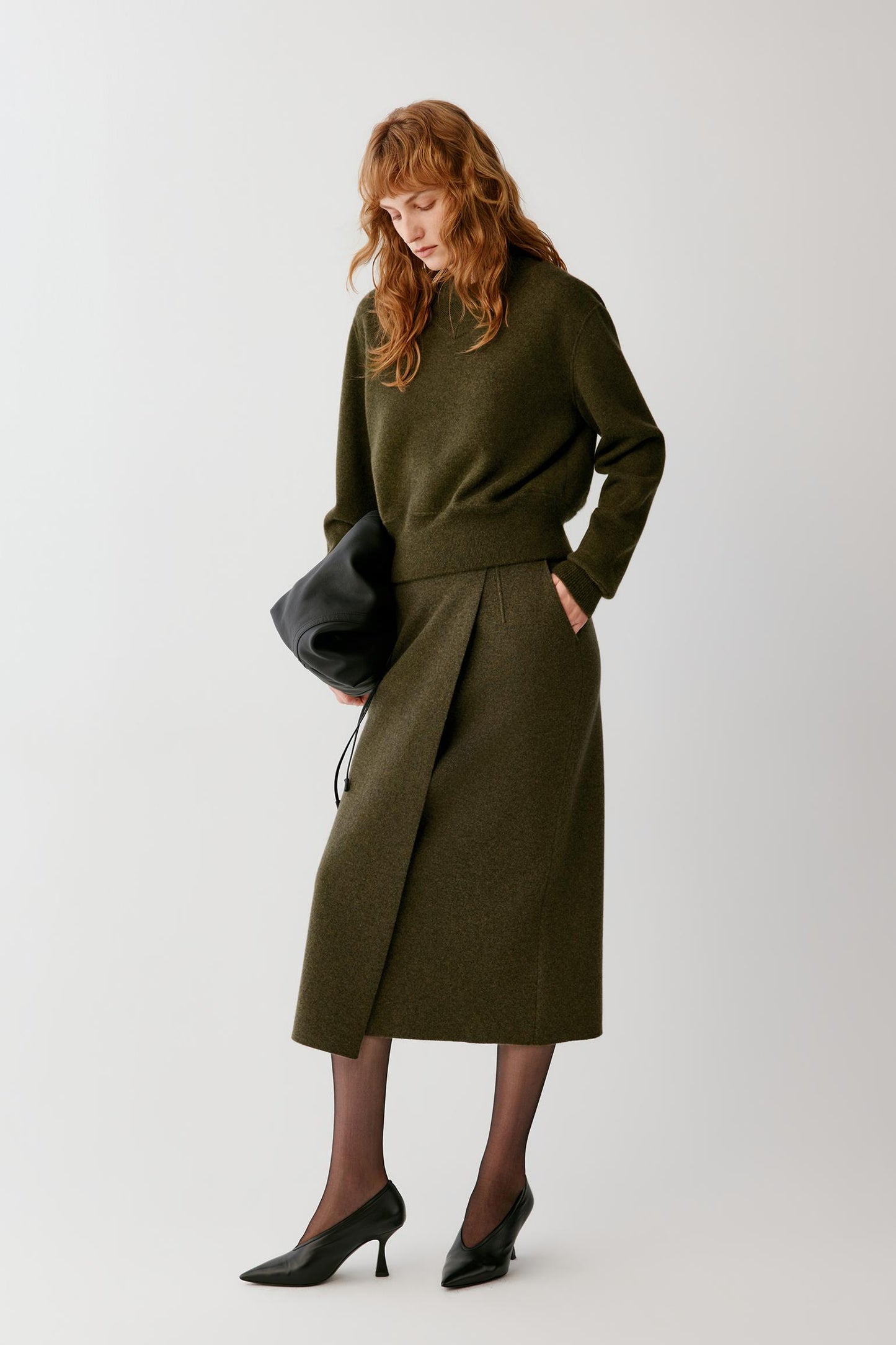 A woman with red hair stands against a light gray studio background wearing an olive green knit sweater and matching pleated midi skirt, holding a black clutch and wearing black pointed heels.