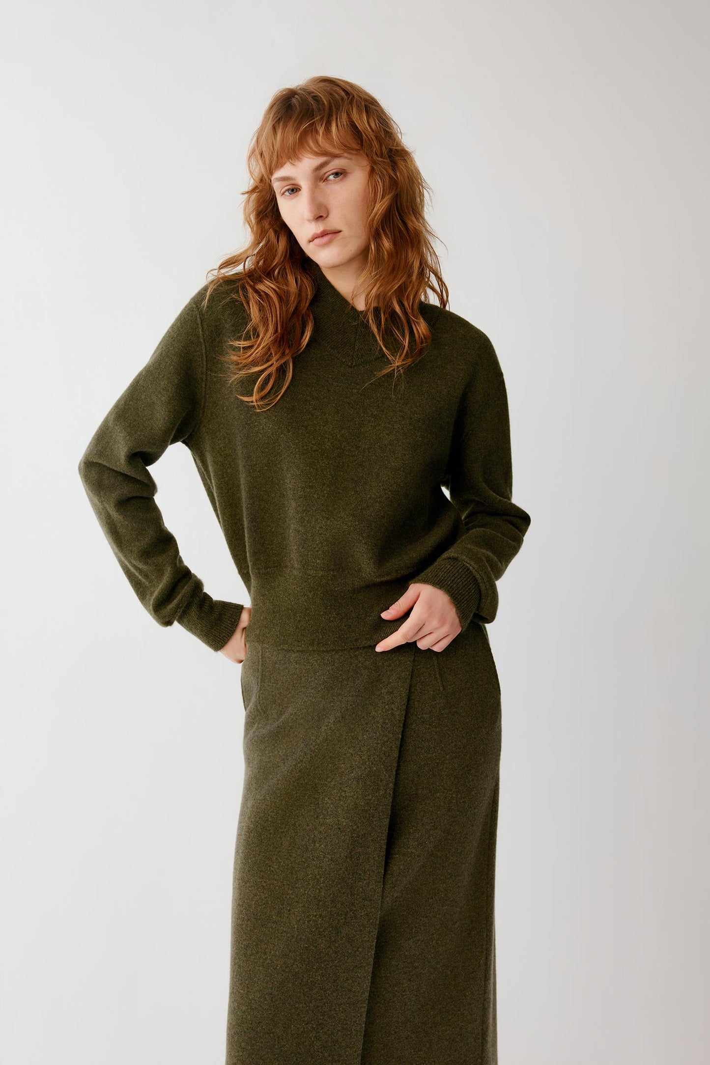 A woman with red hair wearing a matching olive green knit sweater and wide-leg trousers, standing against a light gray studio background.
