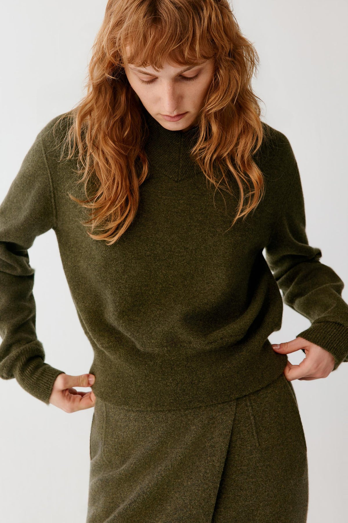 A woman with long wavy auburn hair wearing an olive green knit mock-neck sweater and matching trousers, posing against a light gray studio background.