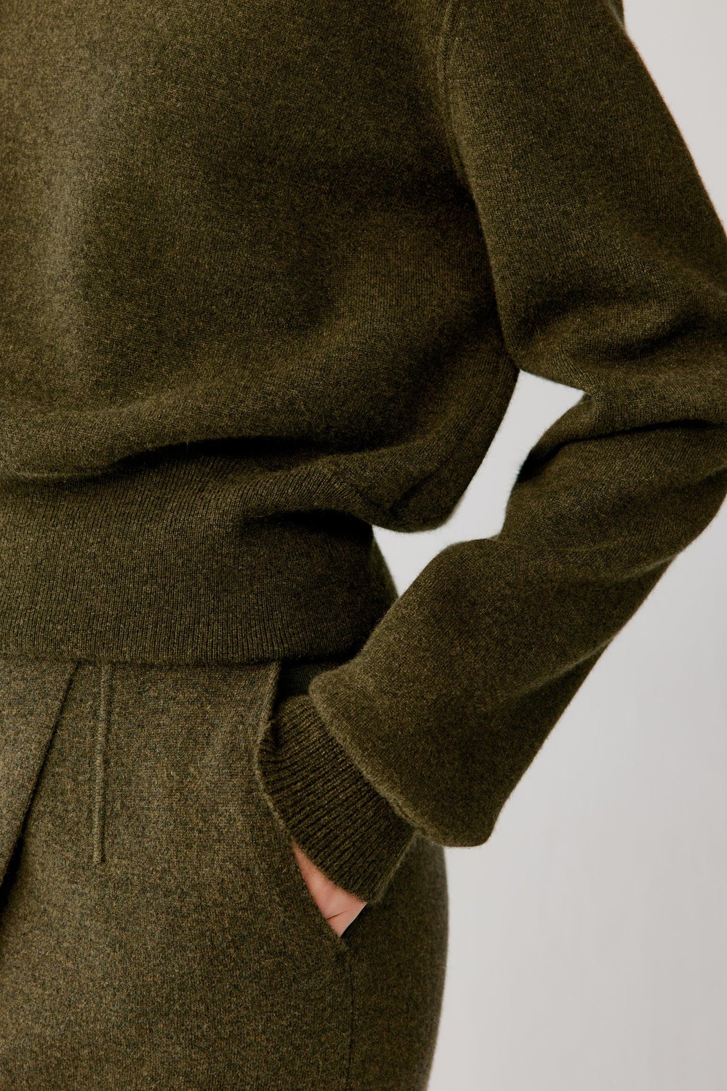 Close-up of a person wearing an olive green knit sweater and matching knit bottoms, with one hand in a pocket against a plain light background.