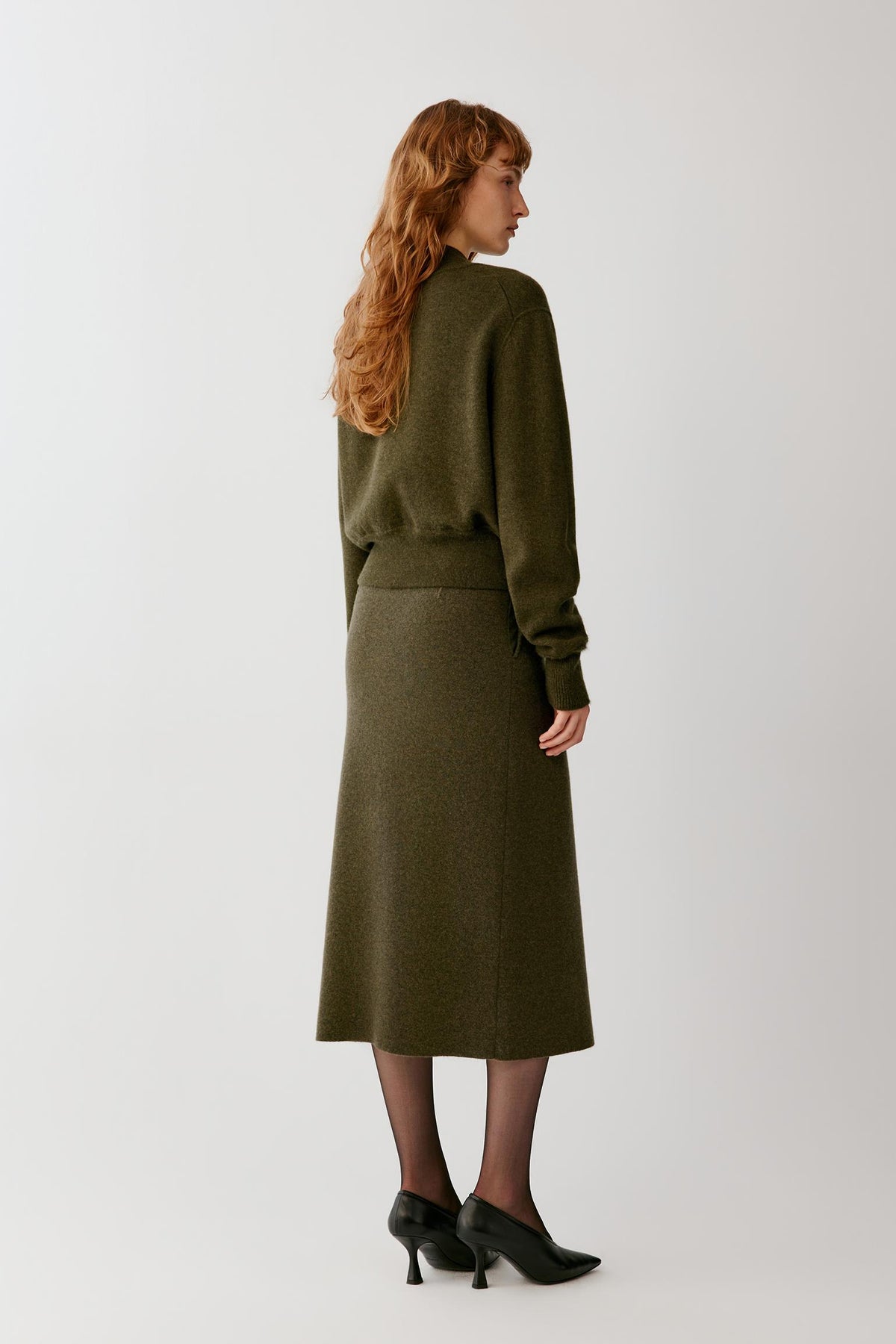 A woman with long wavy red hair is shown from the back wearing an olive green knit sweater and matching midi skirt, paired with black tights and black pointed-toe heels, against a light gray studio background.