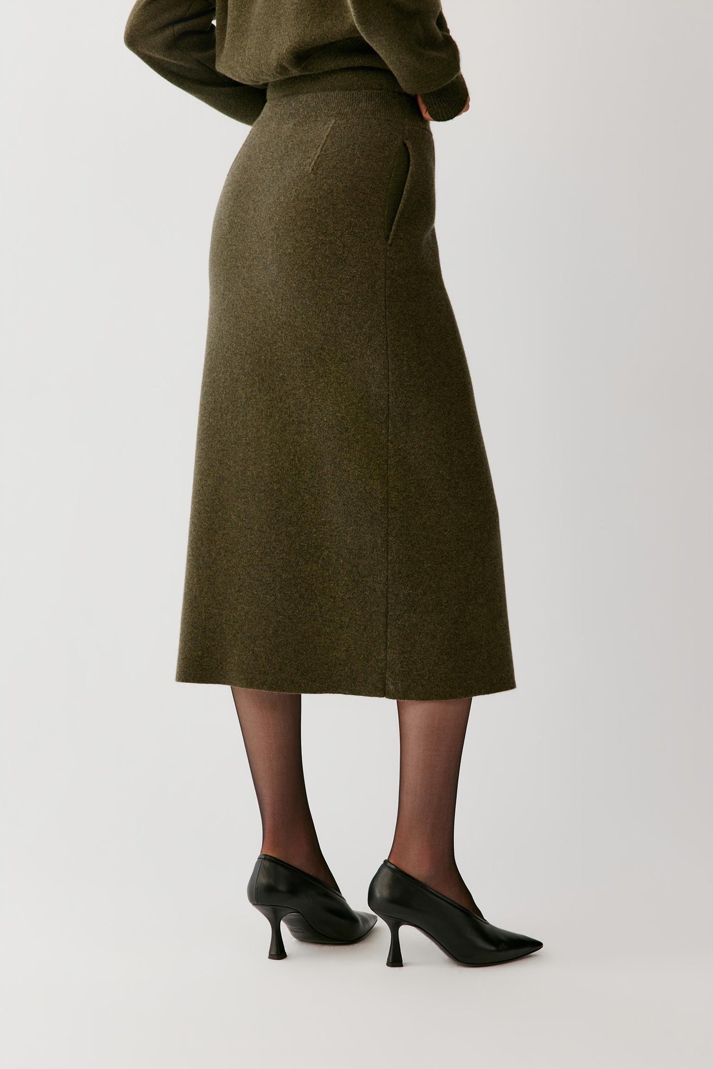 A model shown from the back wearing a dark olive green wool skirt with side pockets, paired with black pointed-toe kitten heel pumps and sheer black tights, against a plain light gray studio background.