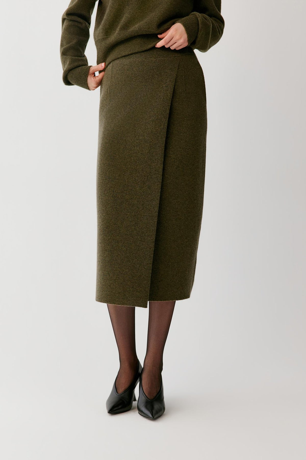 A model is shown from the waist down wearing an olive green knit midi skirt with a wrap-style front, paired with black tights and black pointed-toe heels, against a light gray studio background.