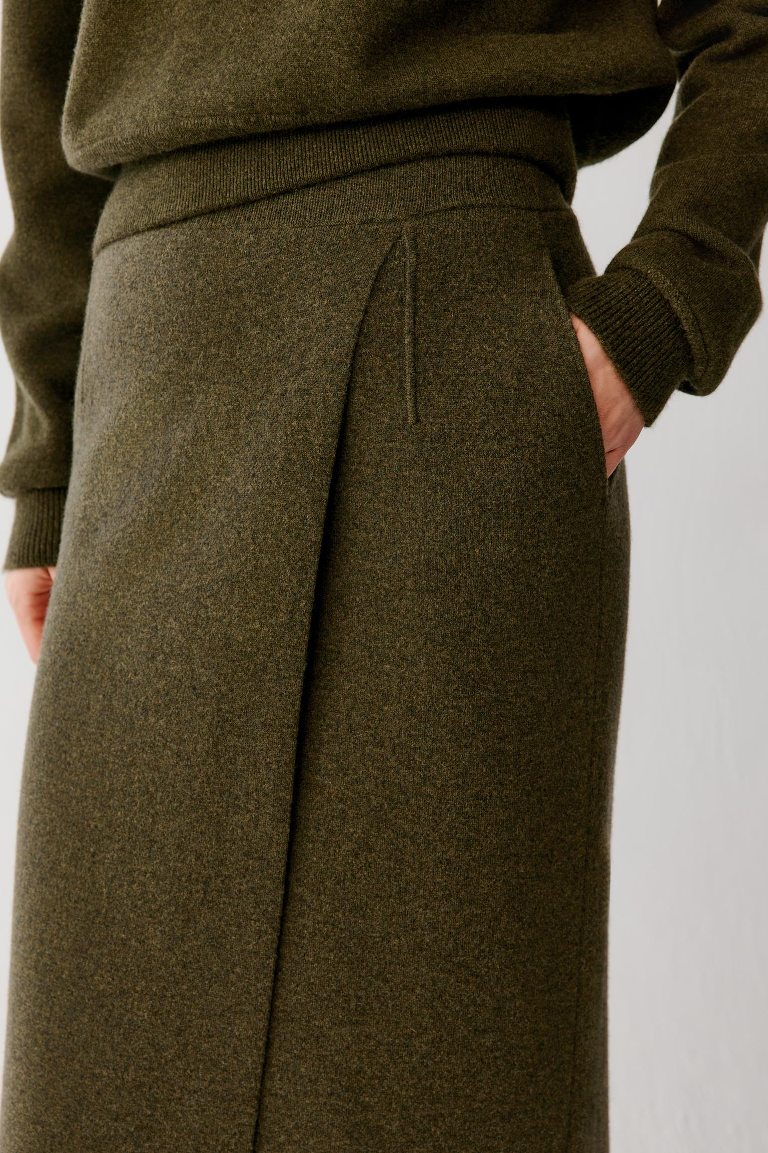 Close-up of a person wearing an olive green knit sweater and matching wide-leg skirt, with one hand in a pocket against a plain light background.