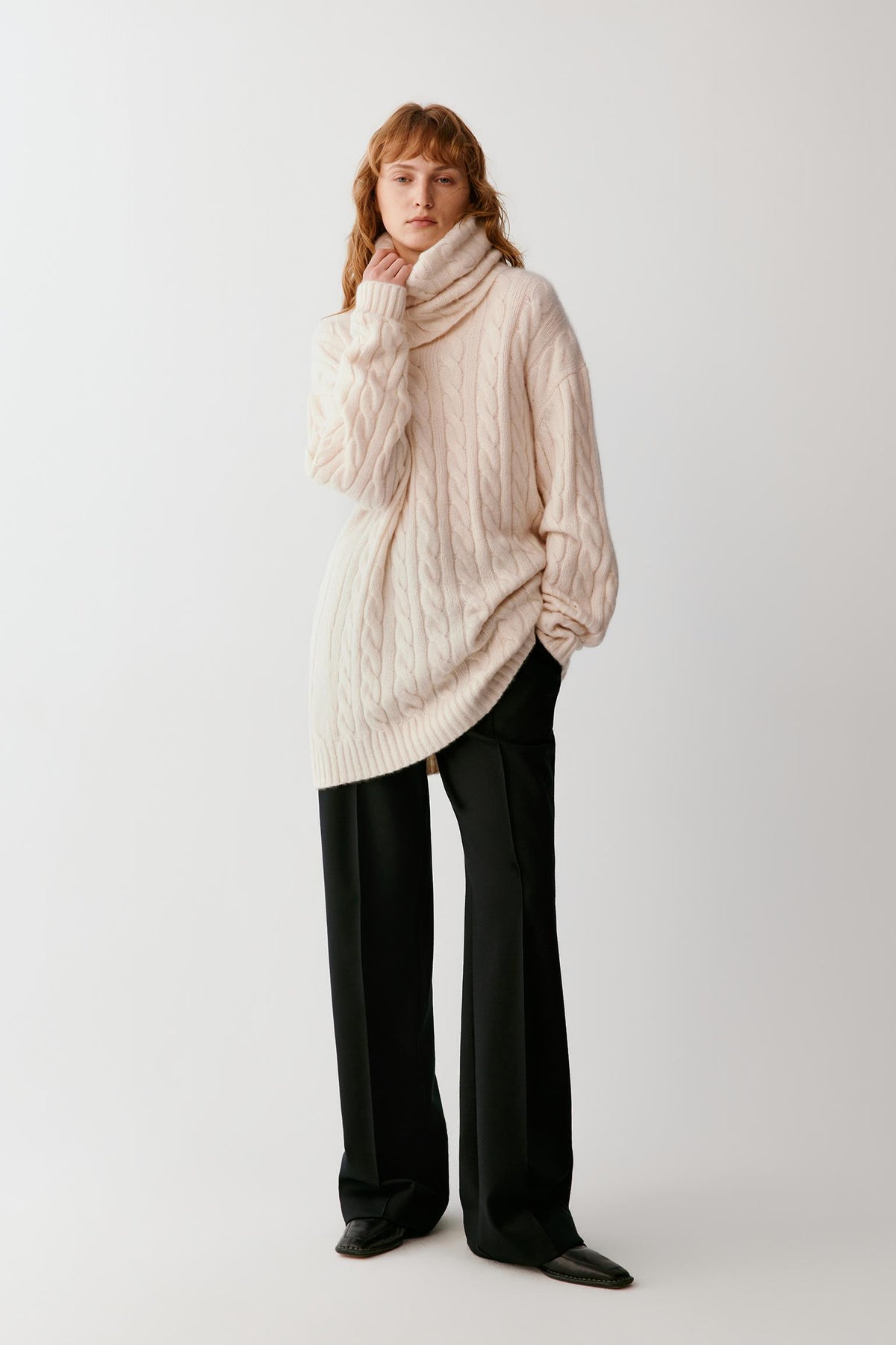 A model wearing a cream-colored cable-knit turtleneck sweater and black wide-leg trousers, standing against a plain light gray studio background.