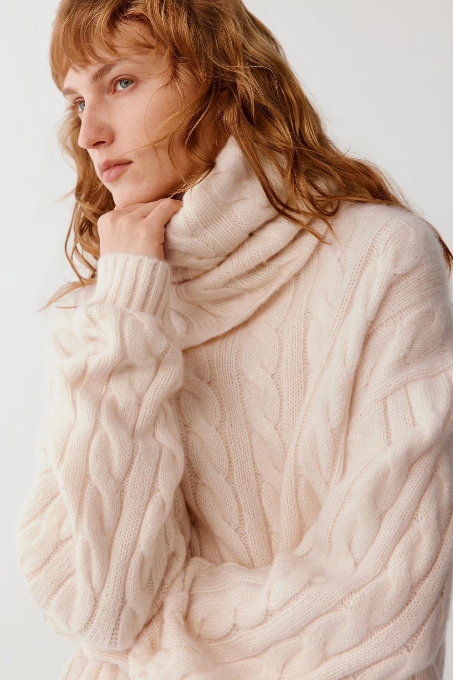A woman wearing a cream-colored cable-knit cowl-neck sweater, posing against a light off-white background.