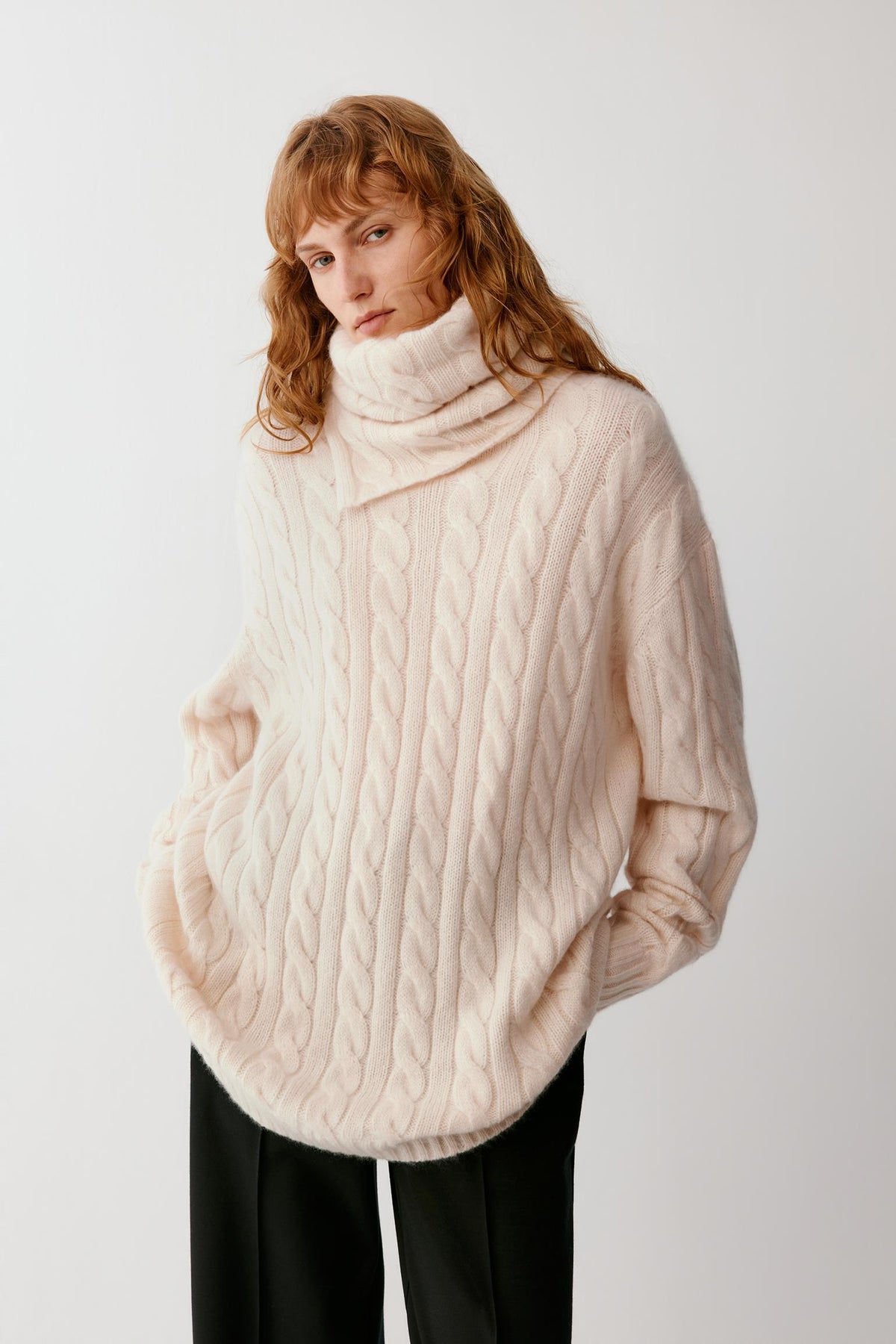 A woman wearing a cream-colored cable-knit turtleneck sweater and black trousers, standing against a plain light gray studio background.