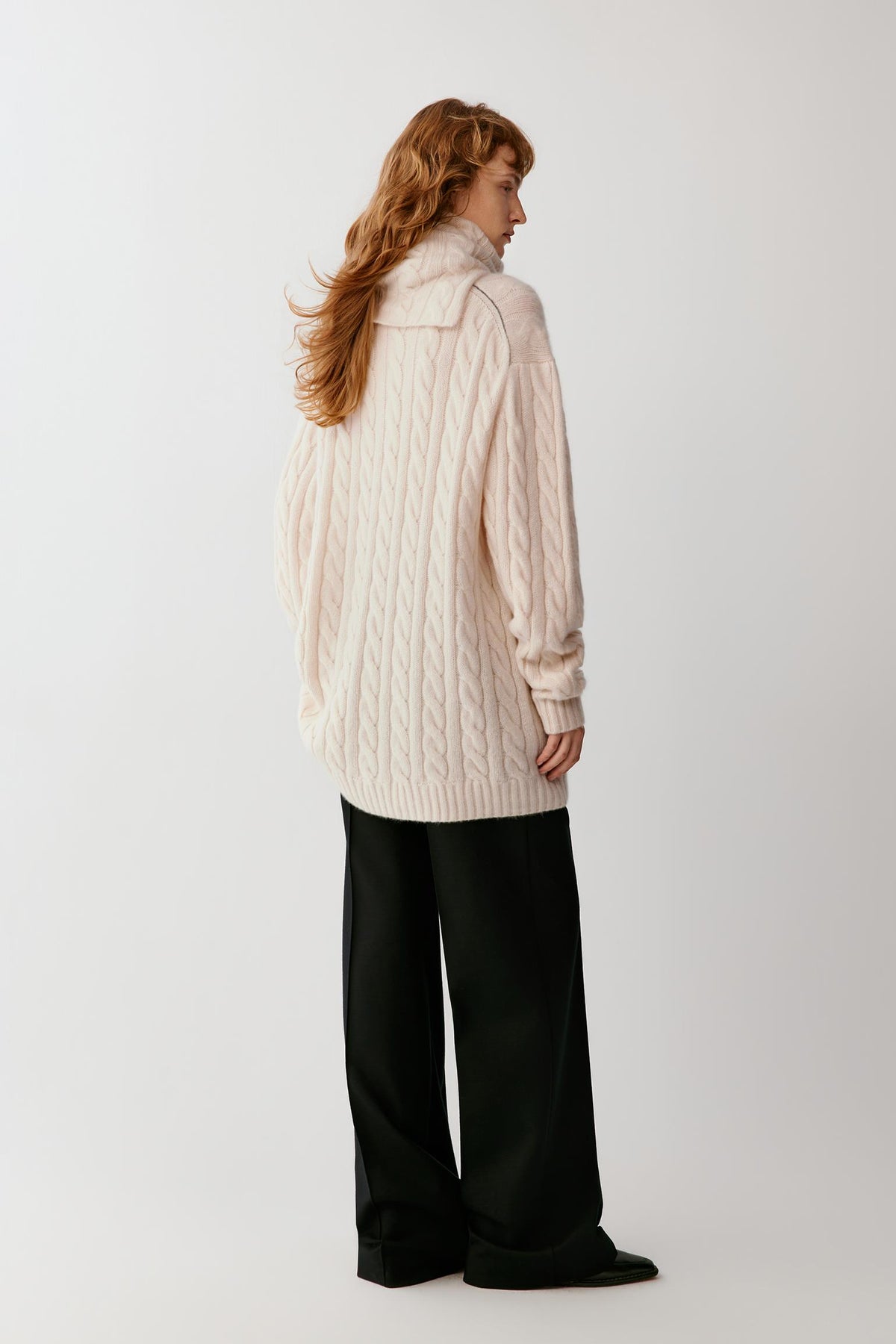 A woman with long red hair is shown from the back wearing an oversized cream cable-knit sweater and black wide-leg trousers, standing against a plain light gray studio background.