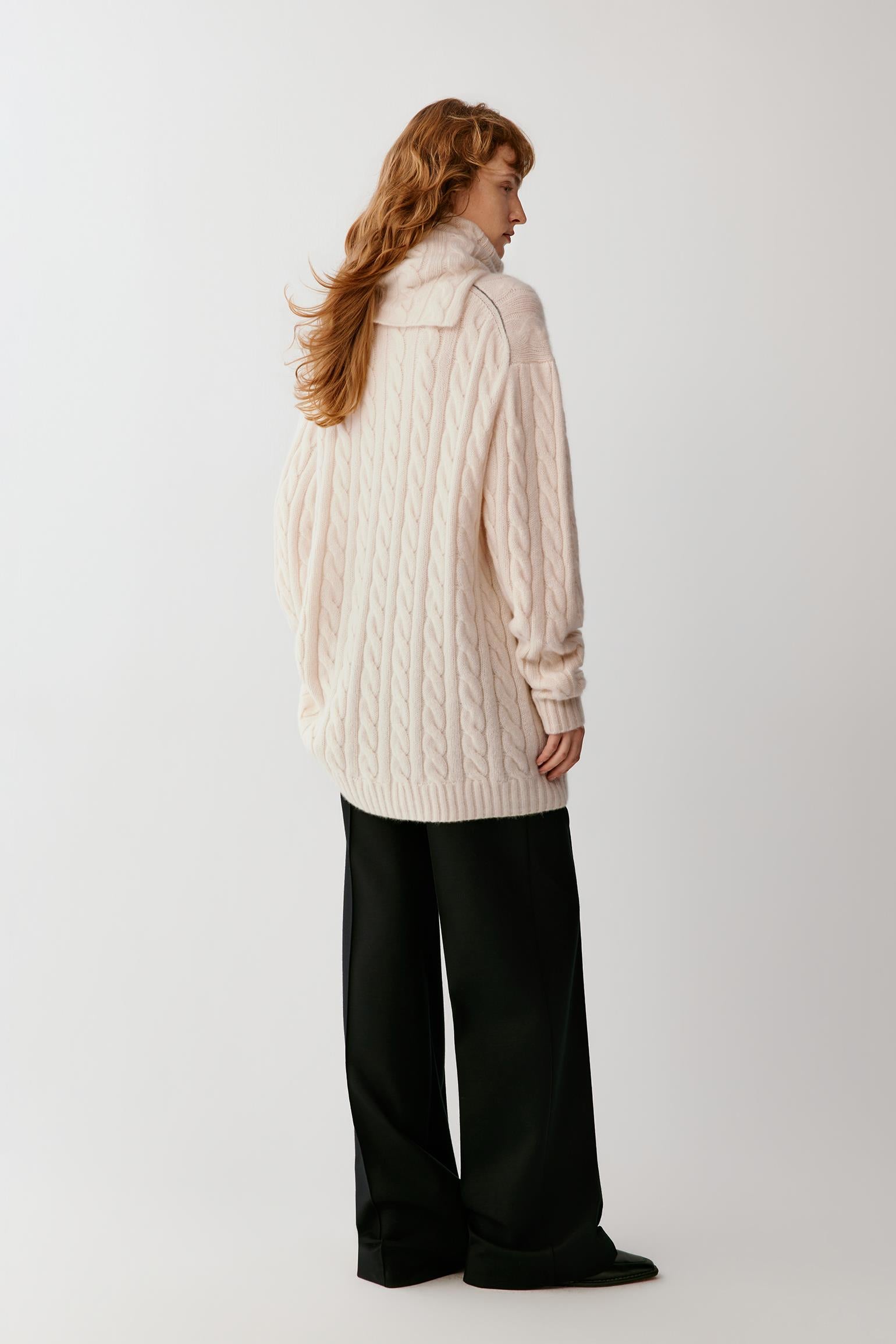 A woman with long red hair is shown from the back wearing an oversized cream cable-knit sweater and black wide-leg trousers, standing against a plain light gray studio background.