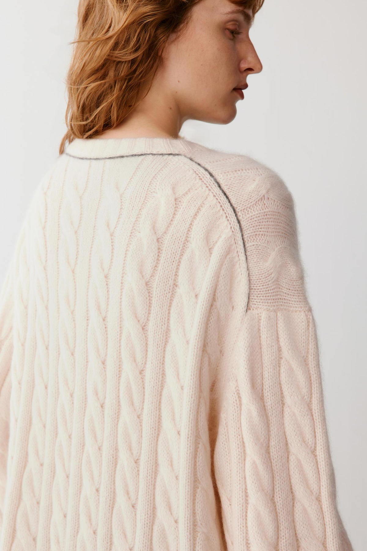 A close-up side view of a person wearing a cream cable-knit sweater against a plain light background.