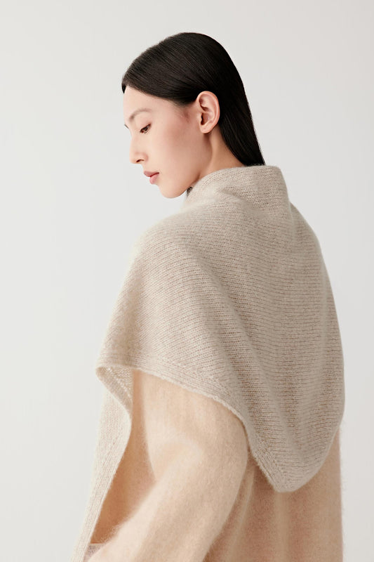 A woman shown from the back wearing a light beige textured knit shawl draped over her shoulders, paired with a soft cream-colored sweater, against a plain light gray background.