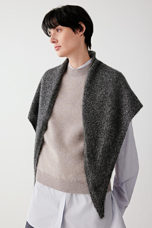 A model wearing a dark gray textured knit poncho draped over a beige crewneck sweater and white shirt, photographed against a clean light gray studio background.