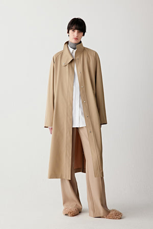 Full-length studio photograph of a woman wearing a long camel trench coat in wool and silk. The coat has a straight, elongated silhouette with a press-stud collar and a concealed front closure. It falls below the calf and is worn over matching wide-leg trousers and light shoes, photographed against a light grey background.