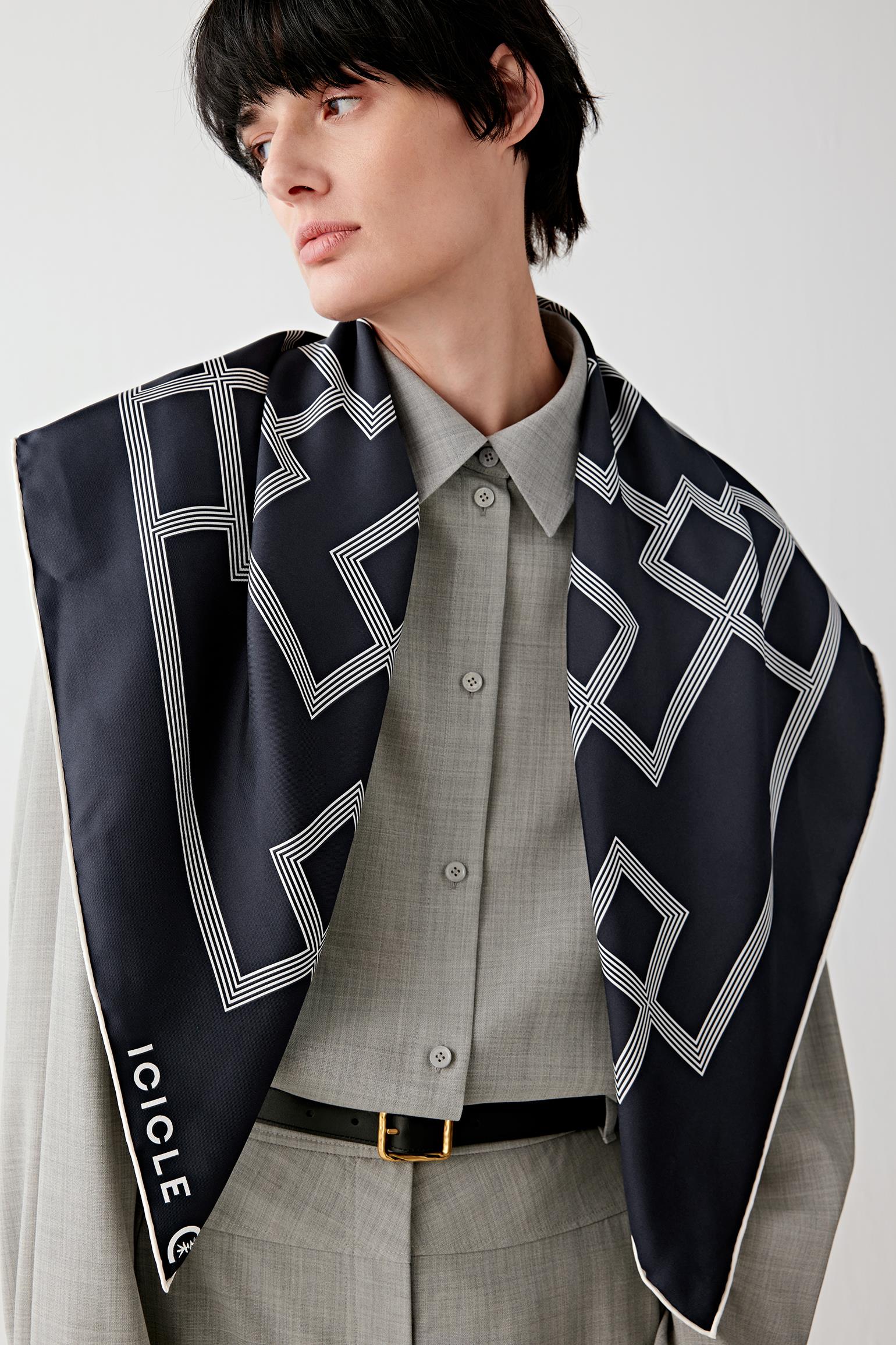 Three-quarter portrait view of a model wearing a navy silk twill scarf with a geometric lattice pattern in contrasting light tones, draped over the shoulders and layered over a shirt and jacket, showing the smooth surface, defined edges and structured drape of the twill weave