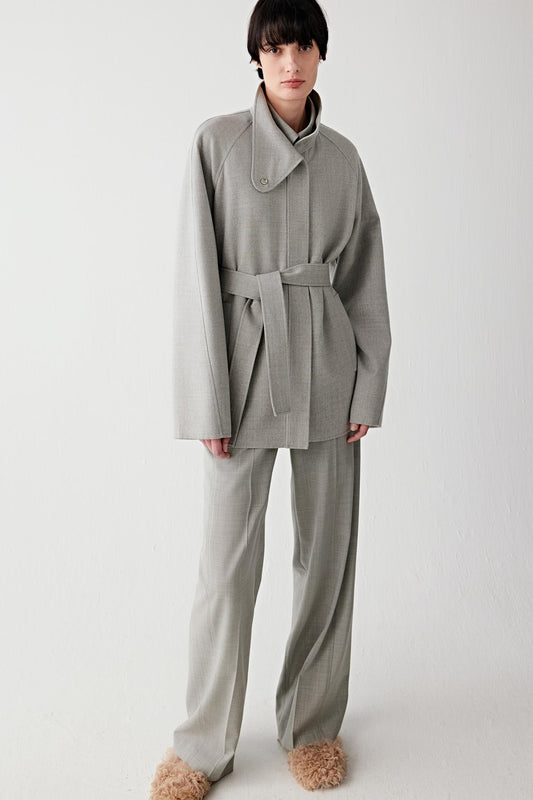 Woman wearing a light grey melange double-faced wool jacket with a relaxed straight cut and self-tie belt at the waist, styled with matching trousers; full-length front view showing the soft structure, clean lines and fluid silhouette.