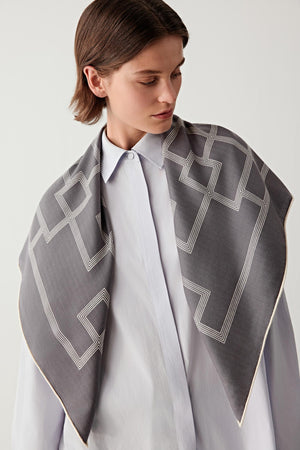 Front view of a model wearing a cashmere and silk scarf draped around the shoulders. The scarf features a soft grey palette with a repeating lattice pattern inspired by traditional Chinese architectural screens.