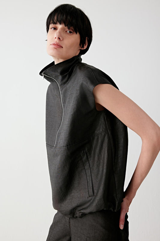 Three-quarter front view of a woman wearing a sleeveless black top in gambiered canton gauze with a high neckline and structured silhouette, showing slight texture and matte finish