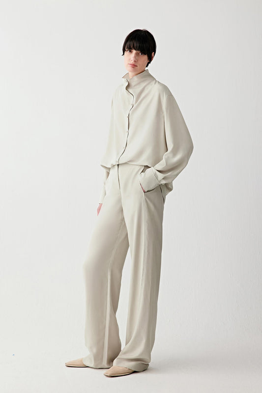 Full-length front view of a woman standing facing forward wearing light beige silk habotai trousers in a mineral shade, styled with a matching blouse, highlighting the fluid straight-leg silhouette, soft drape and smooth, lightly lustrous surface of the fabric