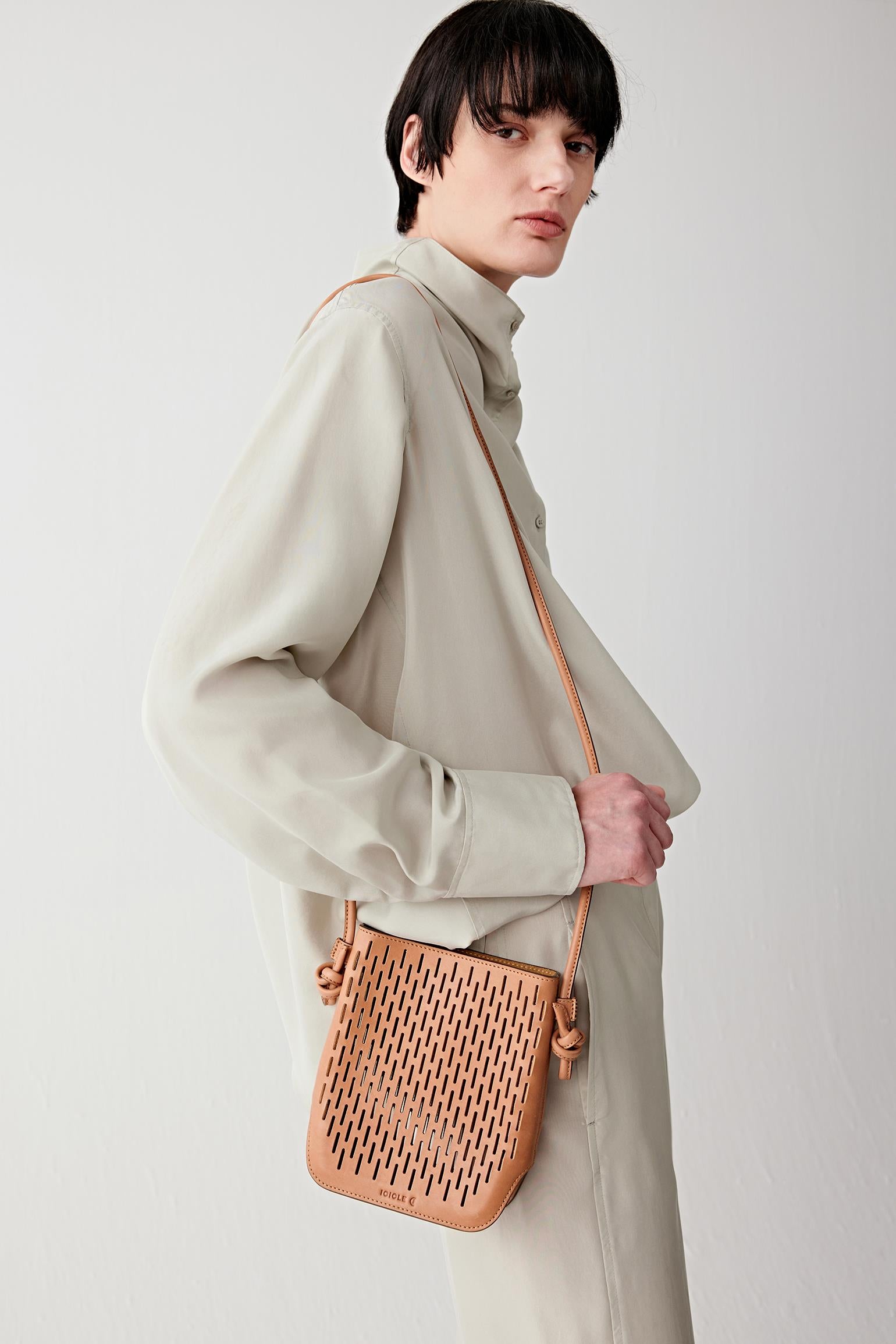 Woman wearing a camel perforated leather crossbody bag with a compact rectangular shape and long adjustable strap, worn across the body, side view highlighting the lightweight construction and clean lines.