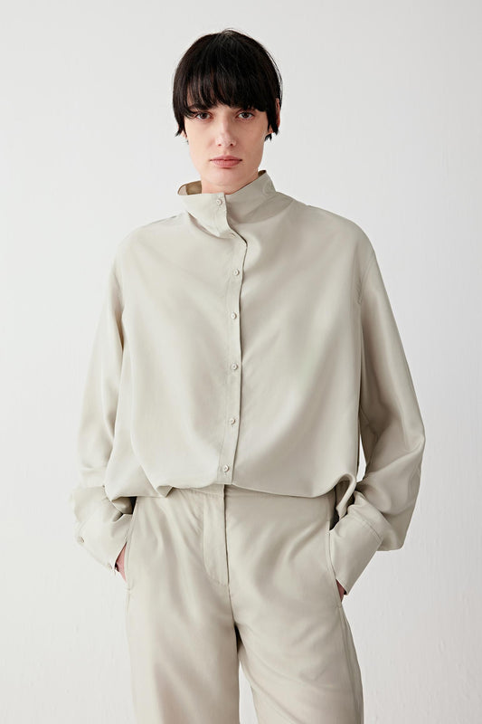 Front view portrait of a woman wearing a light beige silk habotai shirt in a mineral shade, featuring a stand collar and button fastening, highlighting the smooth surface, soft drape and fluid structure of the lightweight silk