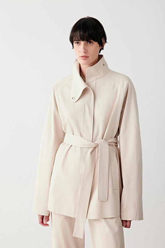 Three-quarter front view of a woman wearing a light beige stretch cotton gabardine jacket with a structured silhouette, high collar and belt, showing smooth surface and tailored lines