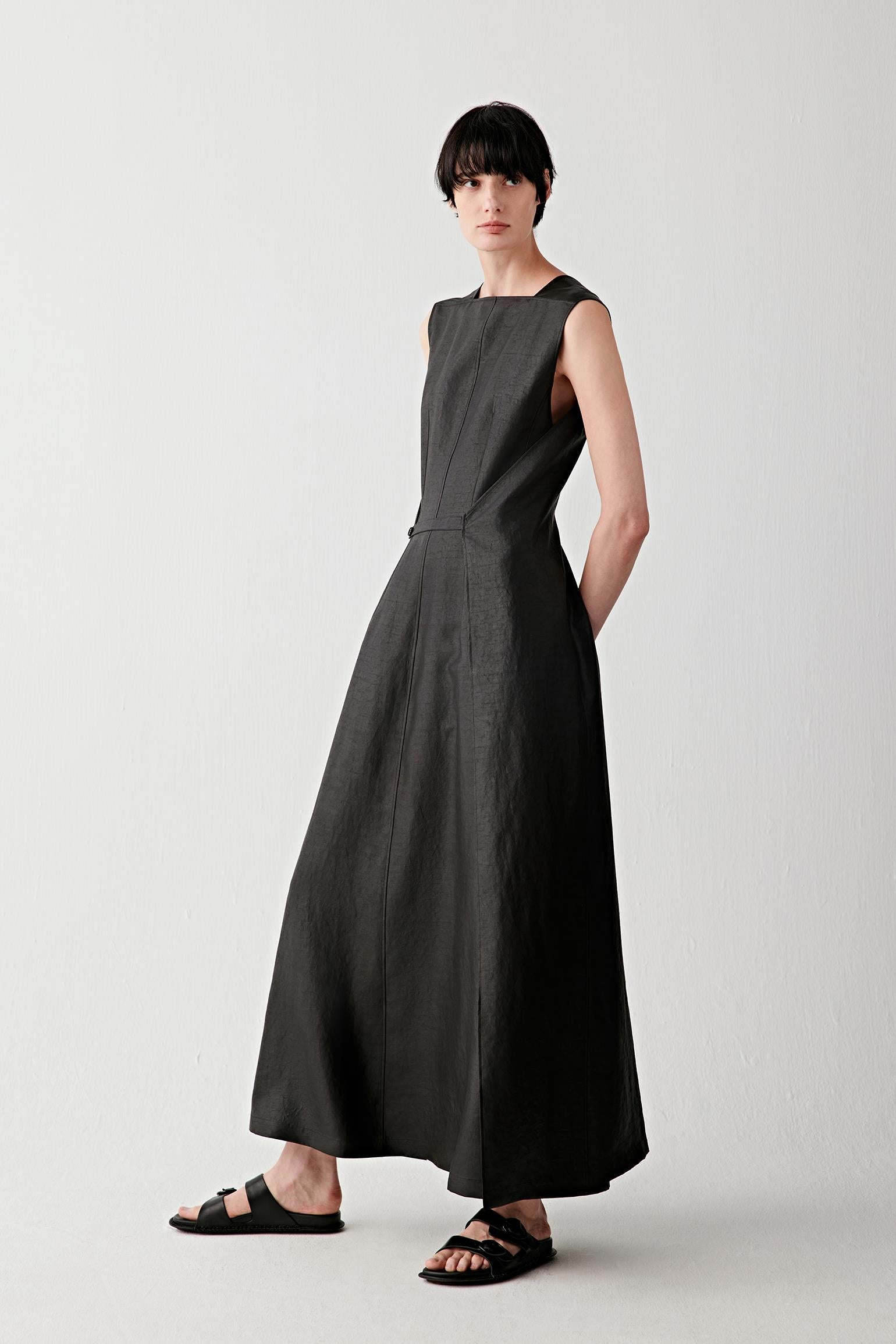 Full-length front view of a woman wearing a sleeveless black dress in gambiered canton gauze with a long fluid silhouette, structured shoulders and soft drape to the ankles